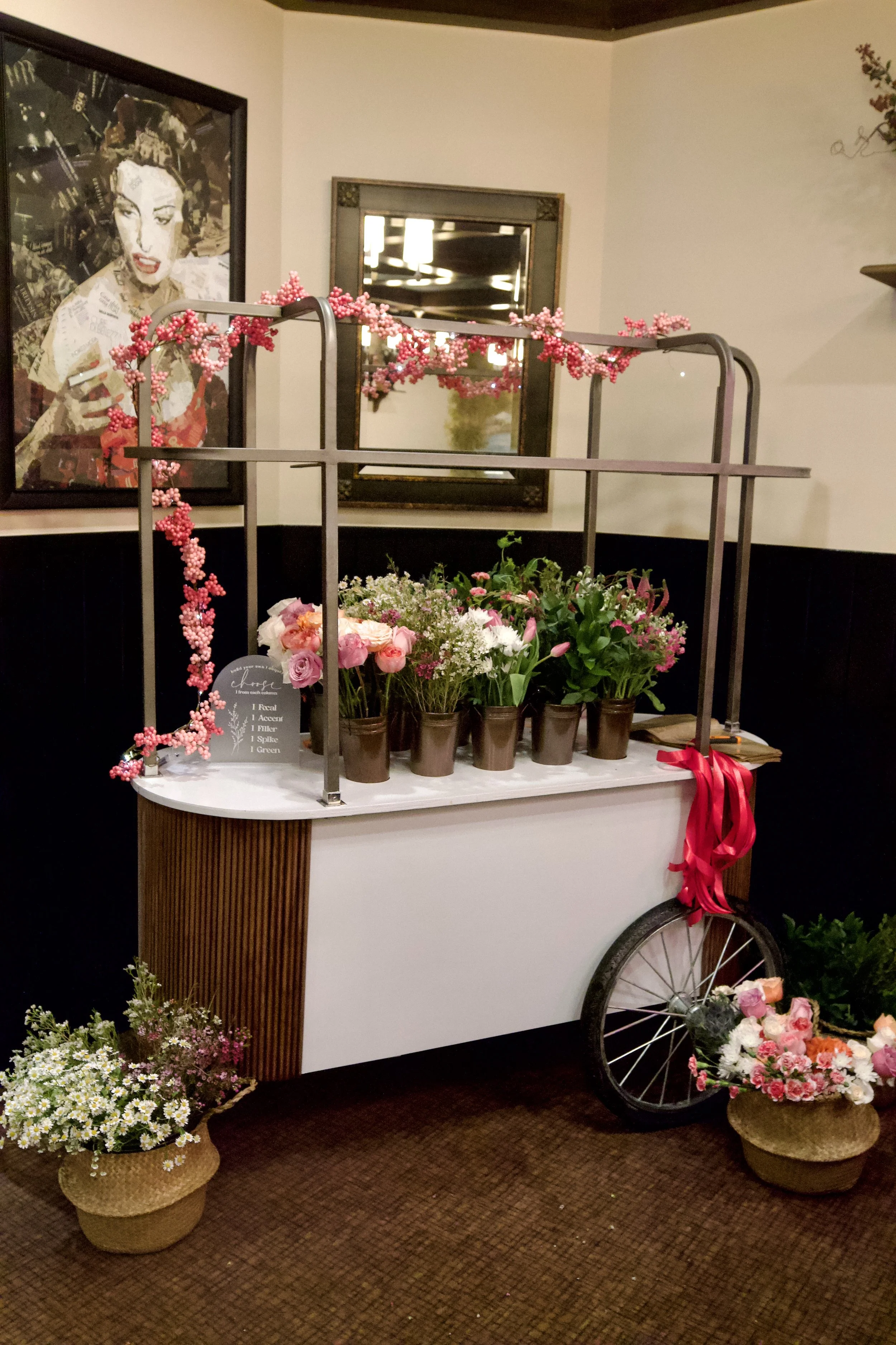 A floral display on a vintage cart with pink and white flowers, pink bead garland, and a ribbon, set in a cozy restaurant with framed art and mirror on the walls at a Bridal Shower in Shelby, MI.