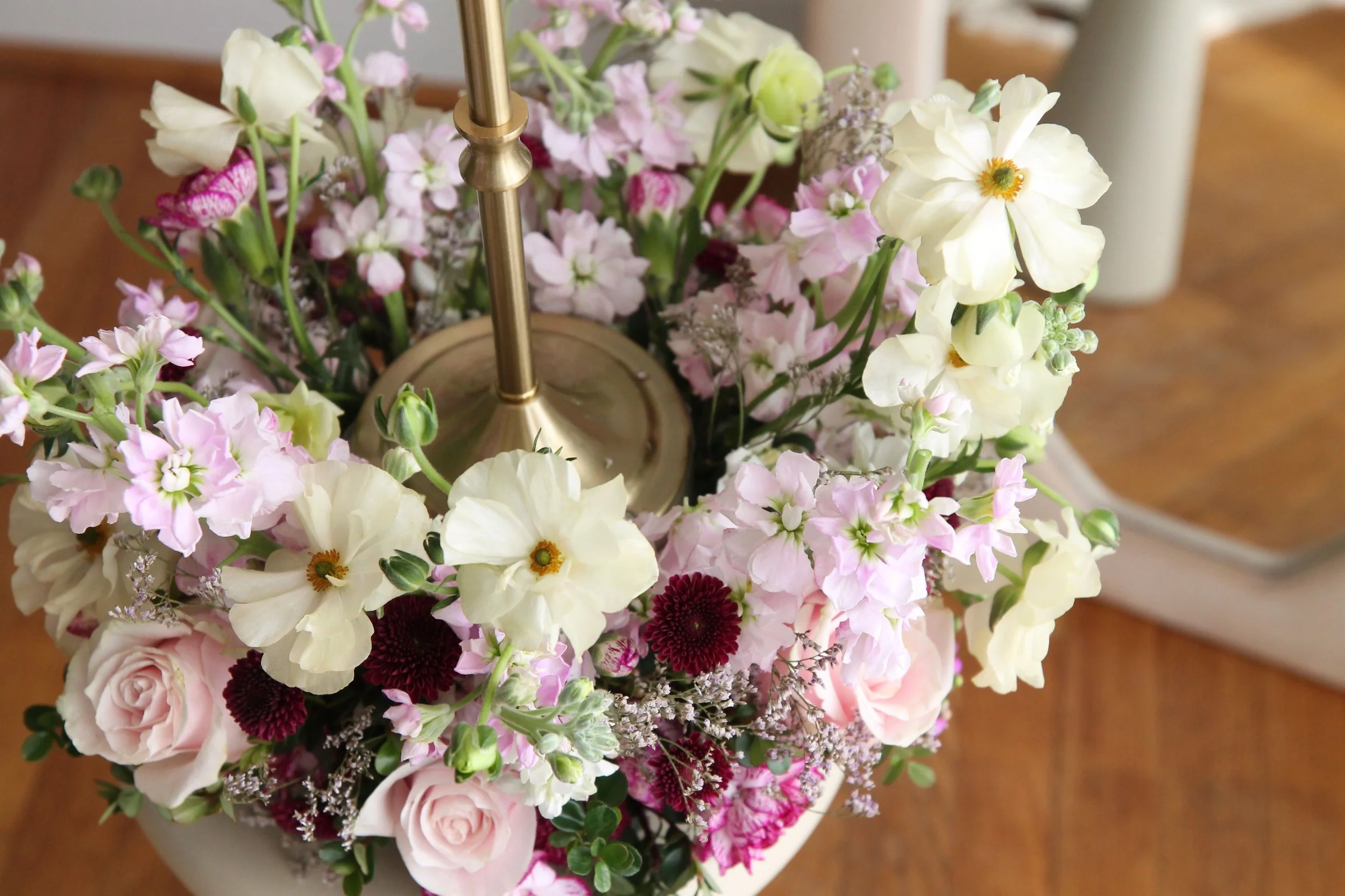 A close-up of a floral arrangement with pink roses, white and pink sweet peas, dark red chrysanthemums, and other small purple and white flowers in a white vase. Part of a brass lamp and a wooden surface are visible.