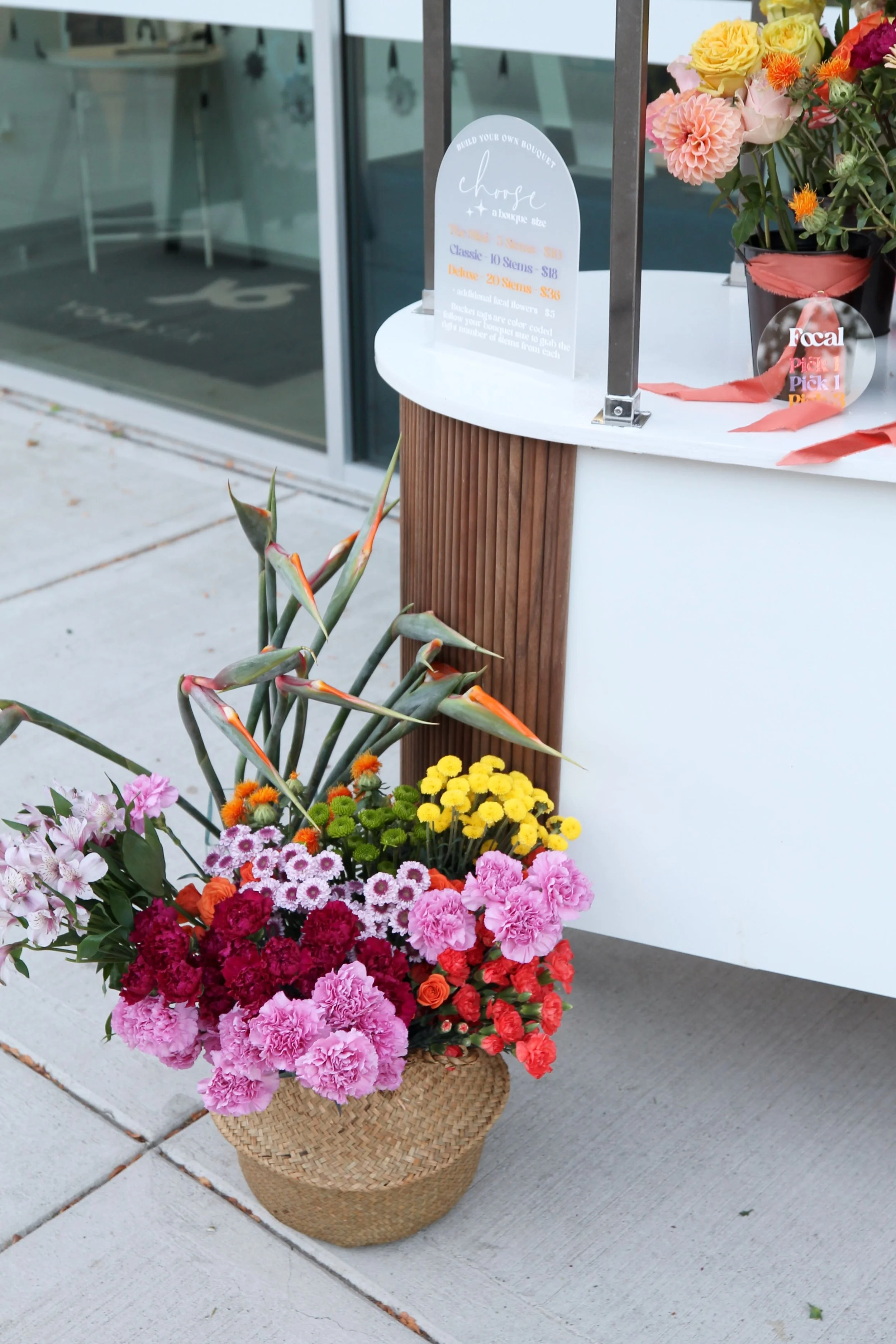 A basket of colorful flowers including pink carnations, red roses, yellow mums, purple daisies, and orange tulips, placed outside near a flower stand with a glass window in the background at a Wedding in Metro Detroit.