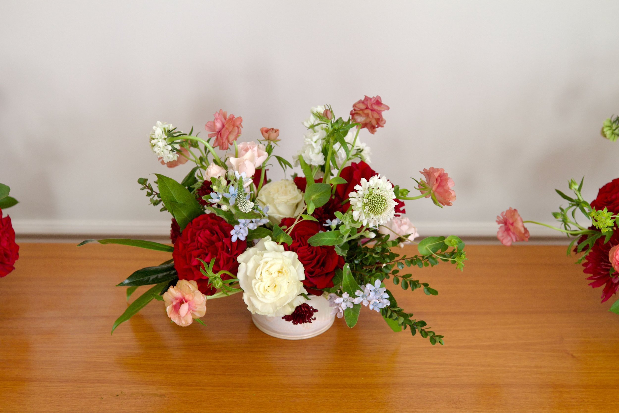 A floral arrangement in a white vase on a wooden table, featuring red, white, pink, and light blue flowers with green leaves.