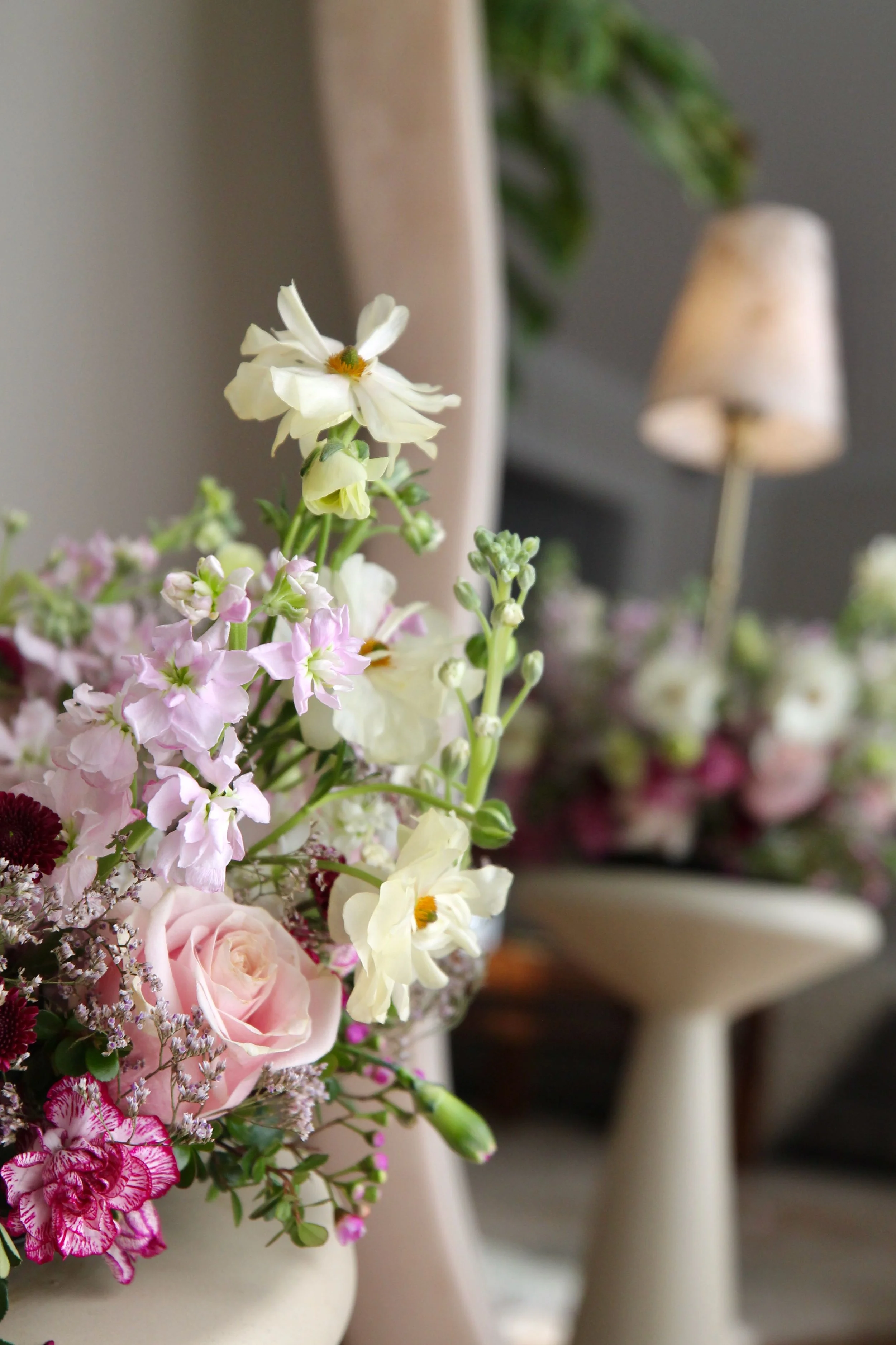 Close-up of a floral arrangement with pink roses, white and pink flowers, and greenery in a white vase. In the background, a blurred table lamp and other floral arrangements are visible.