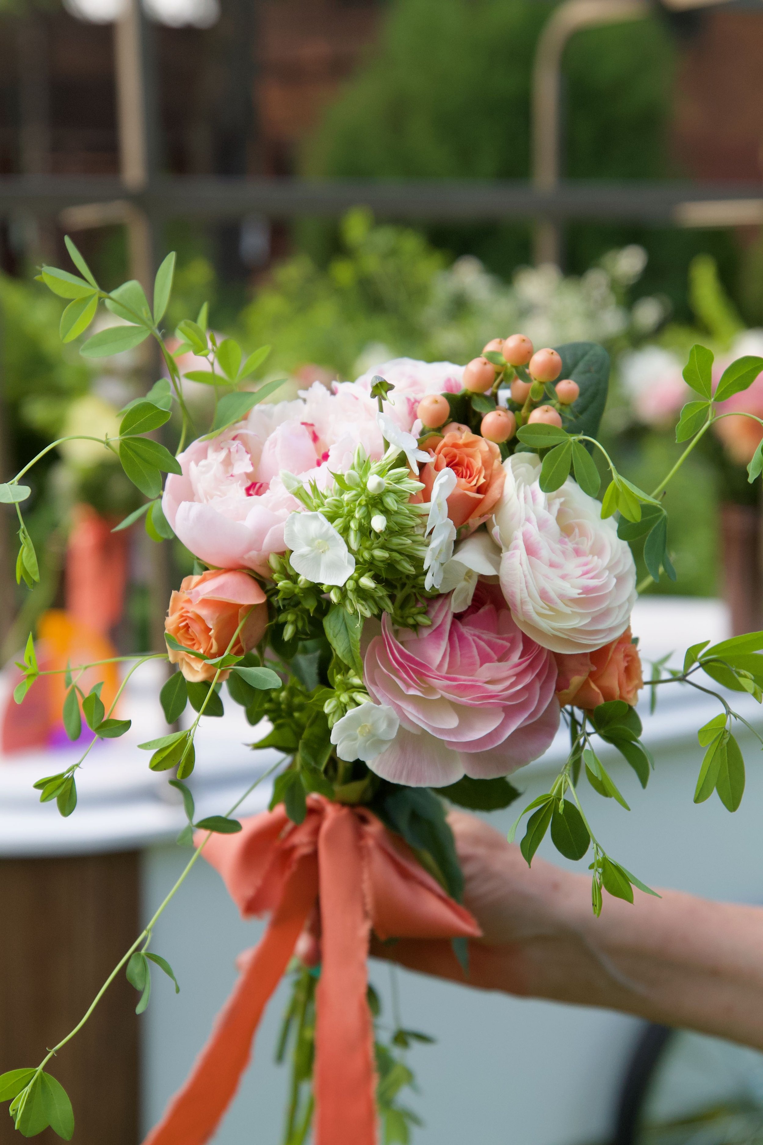 A person's hand holding a bouquet of pink and white flowers, accented with green leaves and tied with an orange ribbon, outdoors with a blurred garden background at a wedding in Birmingham, MI.