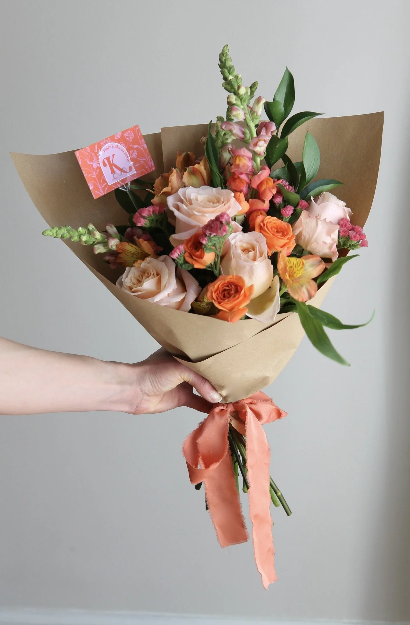 Hand-tied spring bouquet in peach and coral tones featuring garden roses, spray roses, snapdragons, and alstroemeria, wrapped in kraft paper with silk ribbon — handcrafted floral design by Klara’s Bloom Haus in Metro Detroit.