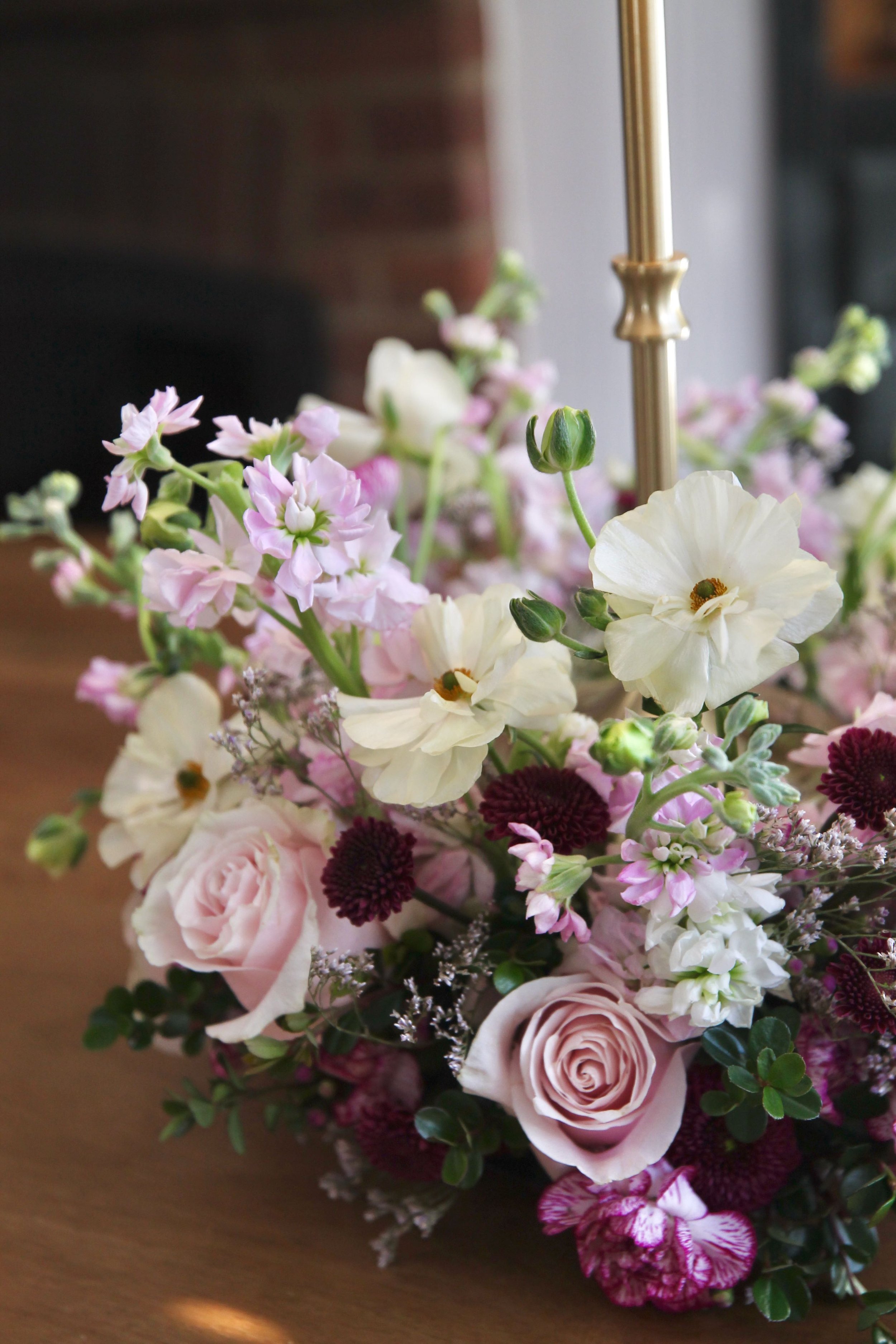 A floral arrangement with pink roses, white snapdragons, dark purple chrysanthemums, and other pink and purple flowers, on a wooden surface with a gold-colored object and a brick wall in the background.