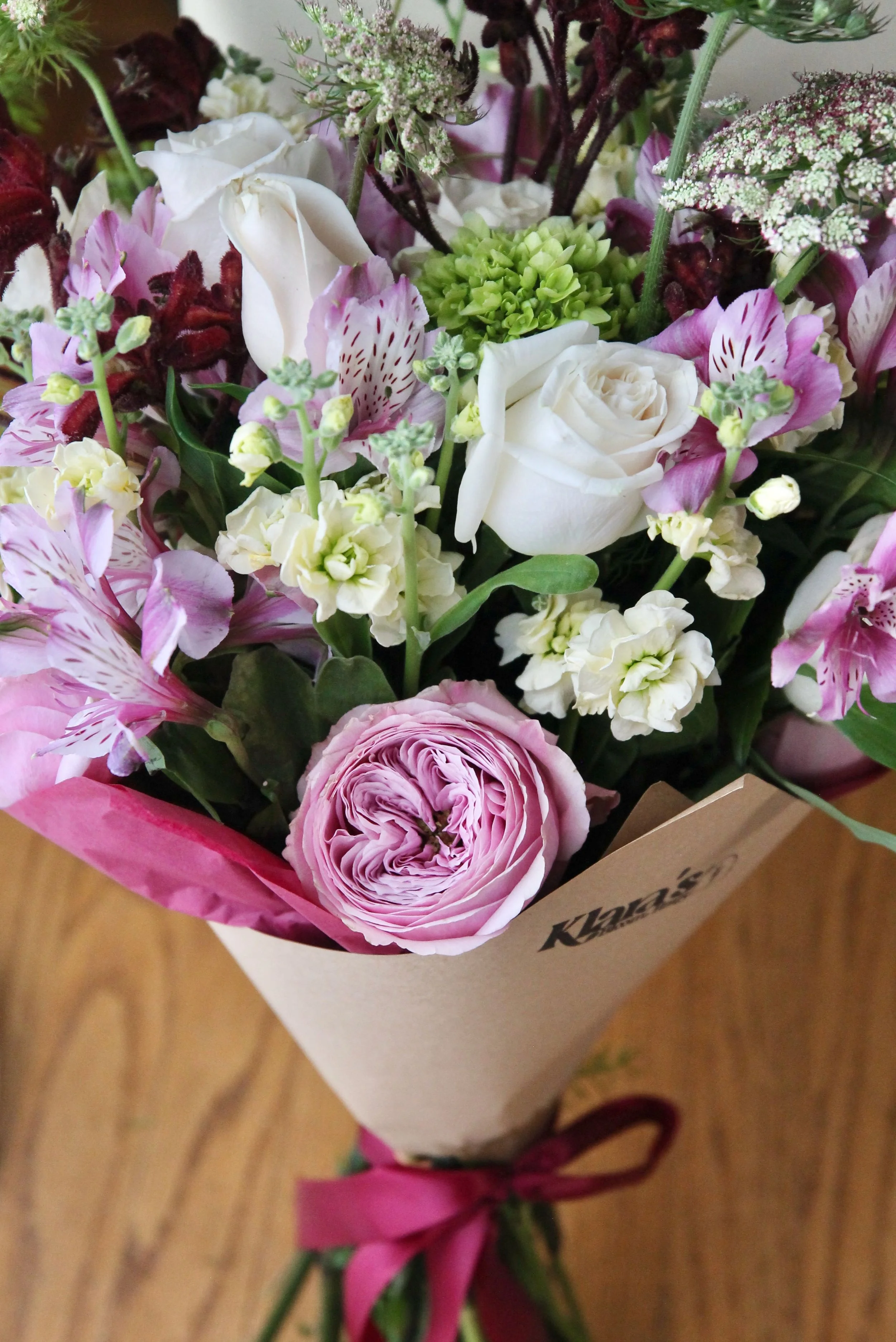 A bouquet of mixed flowers including pink and white roses, purple alstroemeria, green hydrangea, and other small flowers, wrapped in brown paper with a pink ribbon.