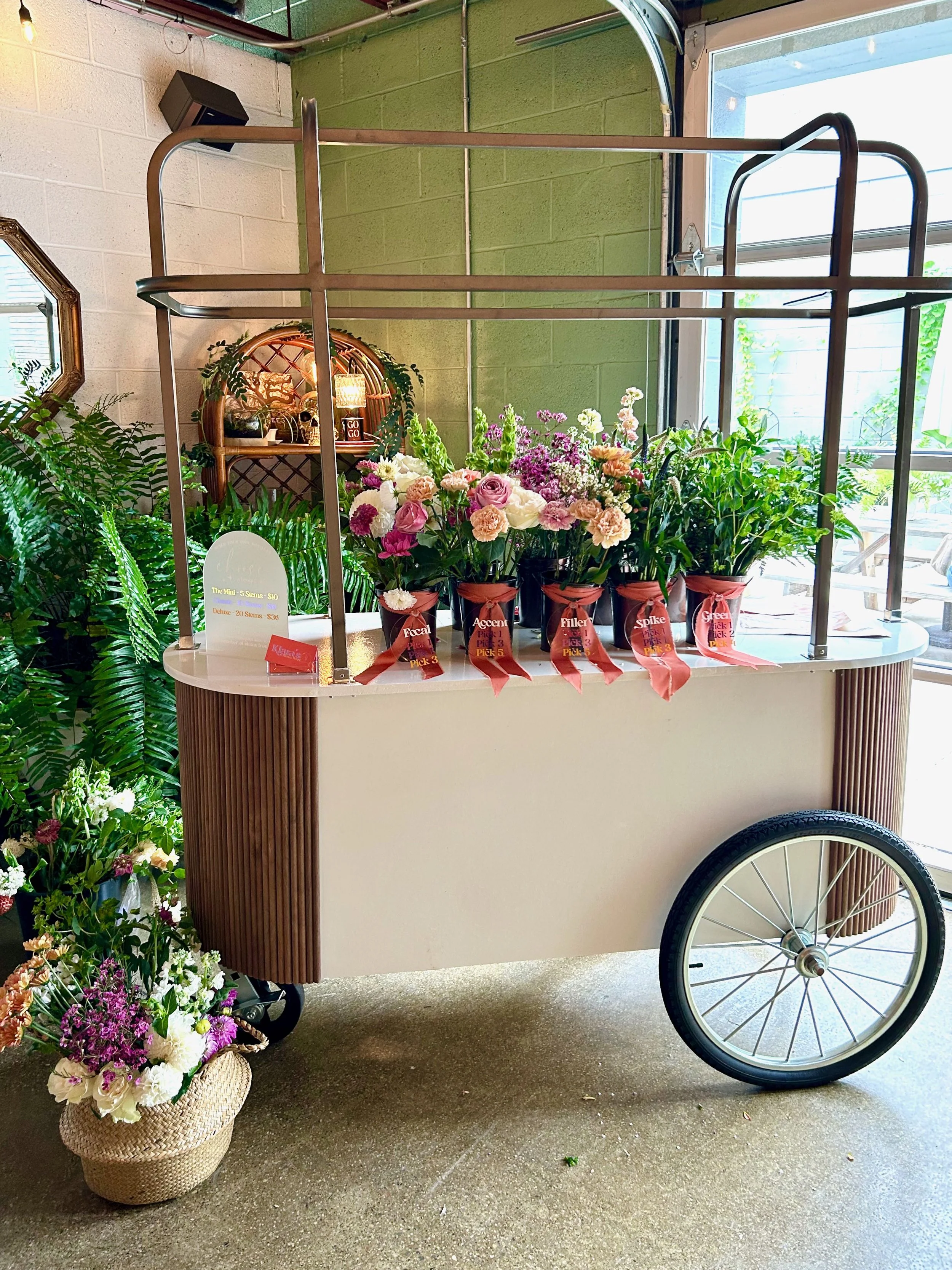 A flower cart filled with potted flowers, mostly pink and white, with labels identifying flower types like focal, accent, filler, spike, and green. There is a basket of flowers on the floor next to the cart. The setting is indoors near a large window, with a green and brick wall in the background.