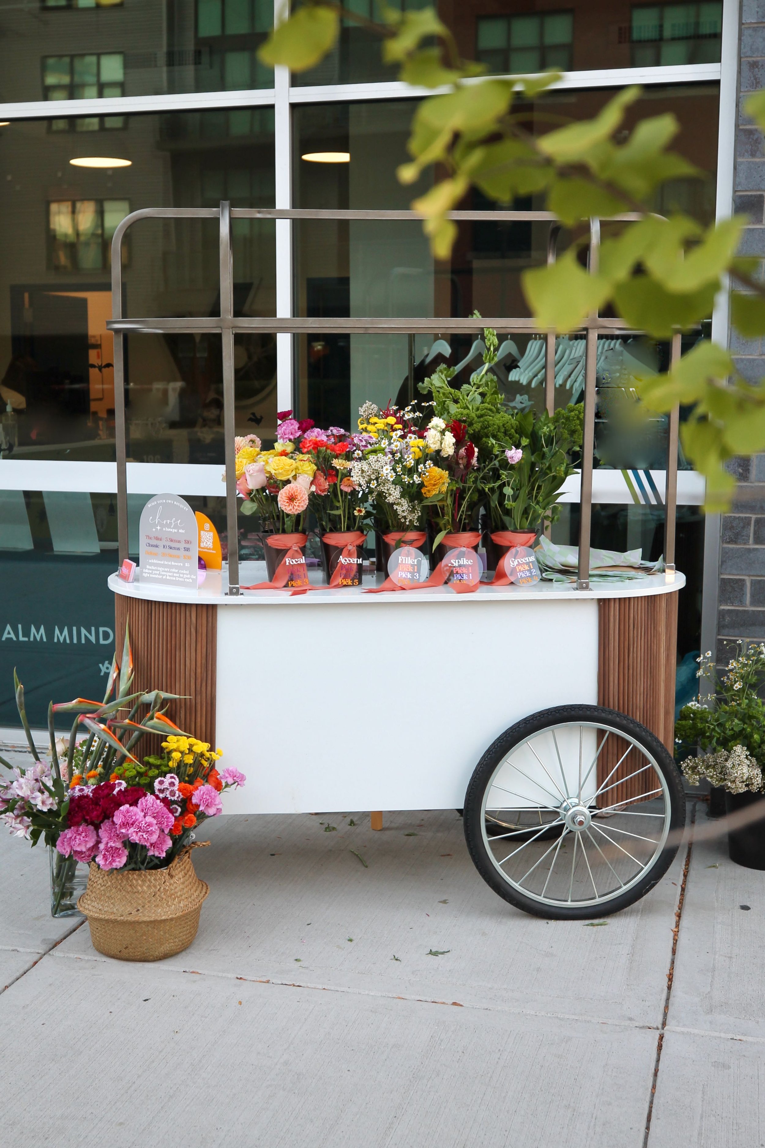 A mobile flower cart with a large wheel, displaying various colorful flowers in small containers. A basket of pink and purple flowers is on the ground next to it, outdoors on a sidewalk in front of a glass building.