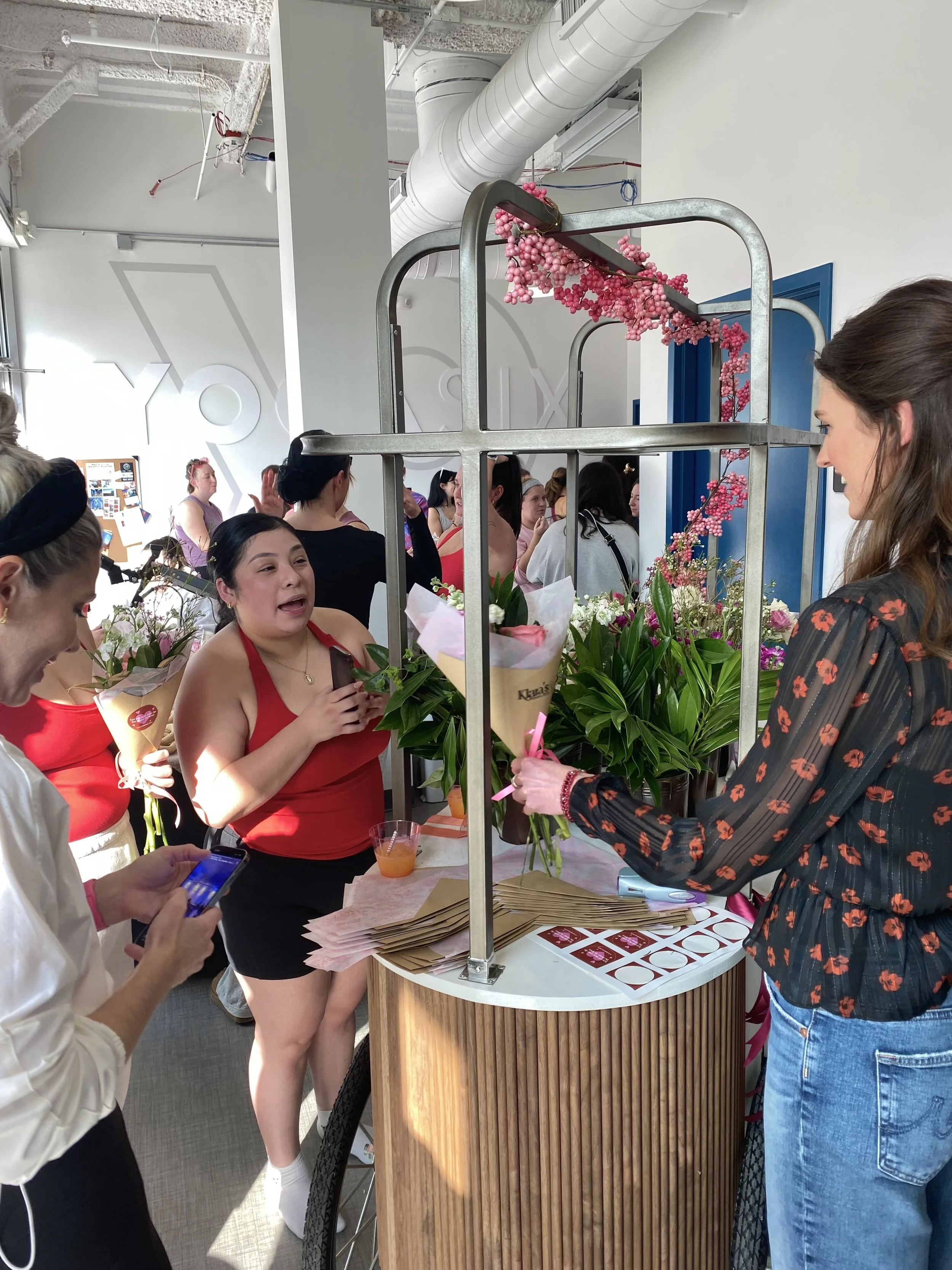 People gathered around a flower stand at an indoor event, with pink and white flowers, some women talking and taking photos, and a woman wearing a black dress with red floral pattern.
