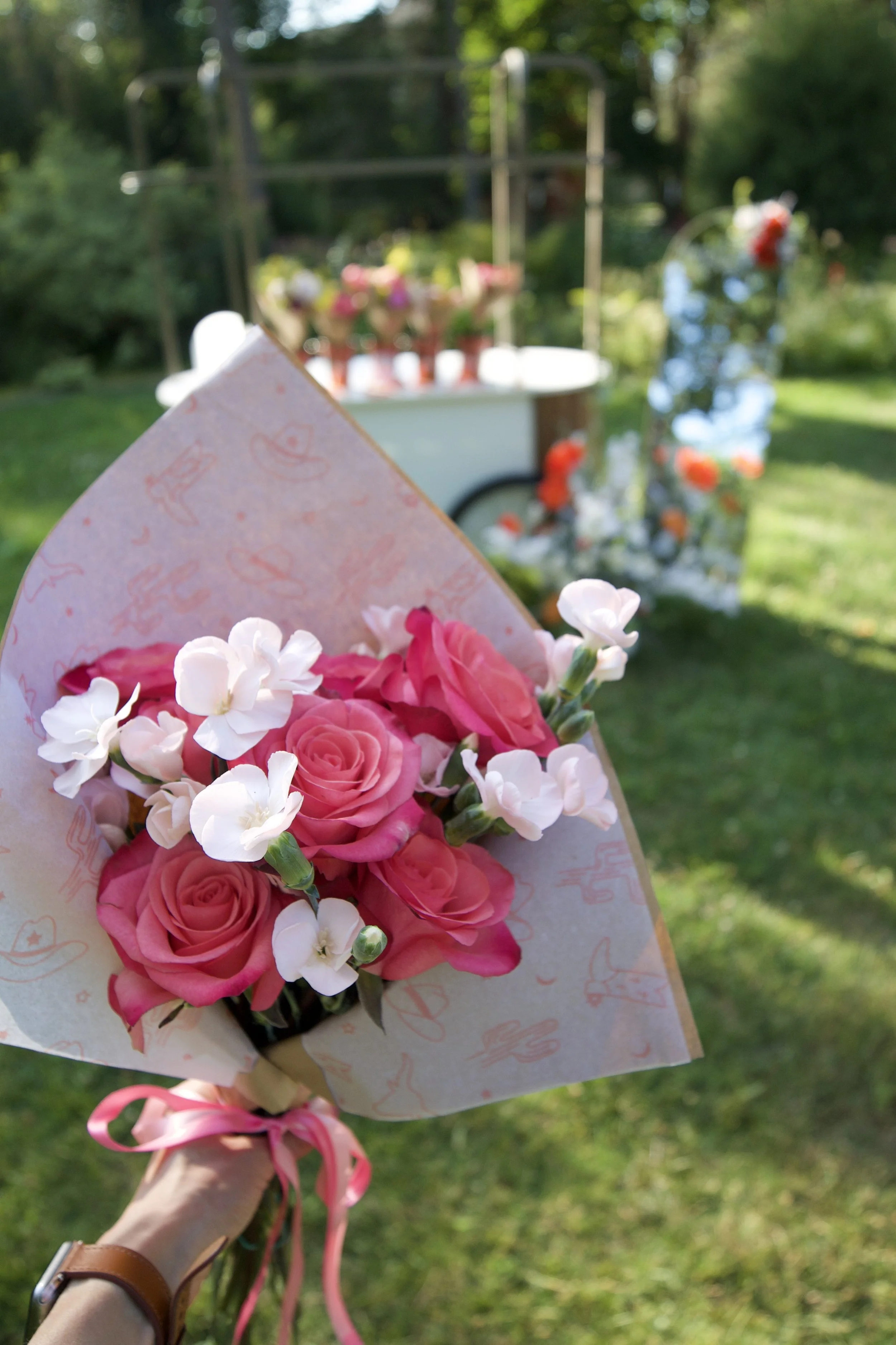 Close-up of a hand holding a small bouquet of pink roses and white flowers, wrapped in patterned paper and tied with a pink ribbon, outdoors in a grassy area with a blurred background of a cart with flowers and greenery.