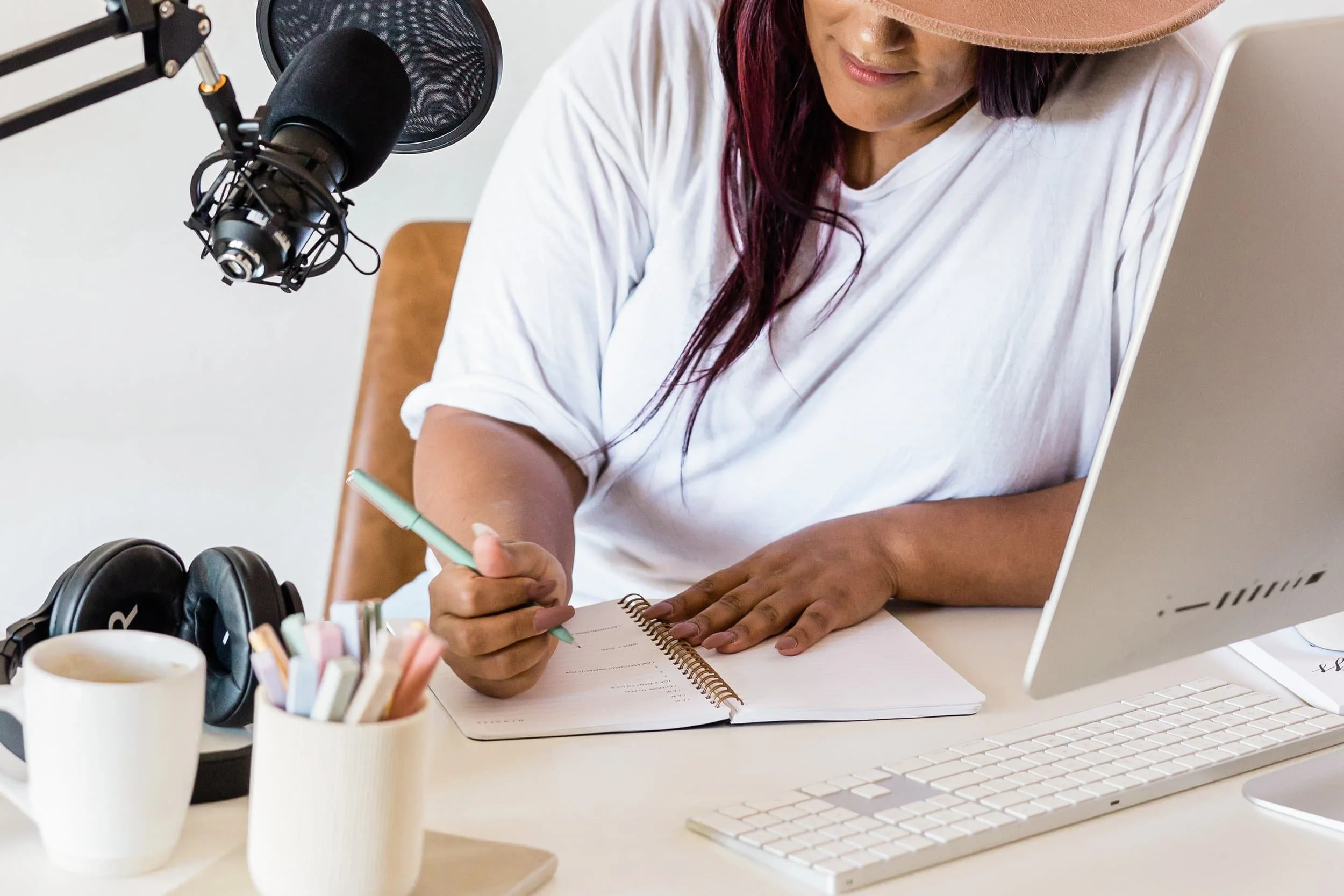 Woman with white shirt and brown hat writing in notebook while at a desk with a computer and microphone.