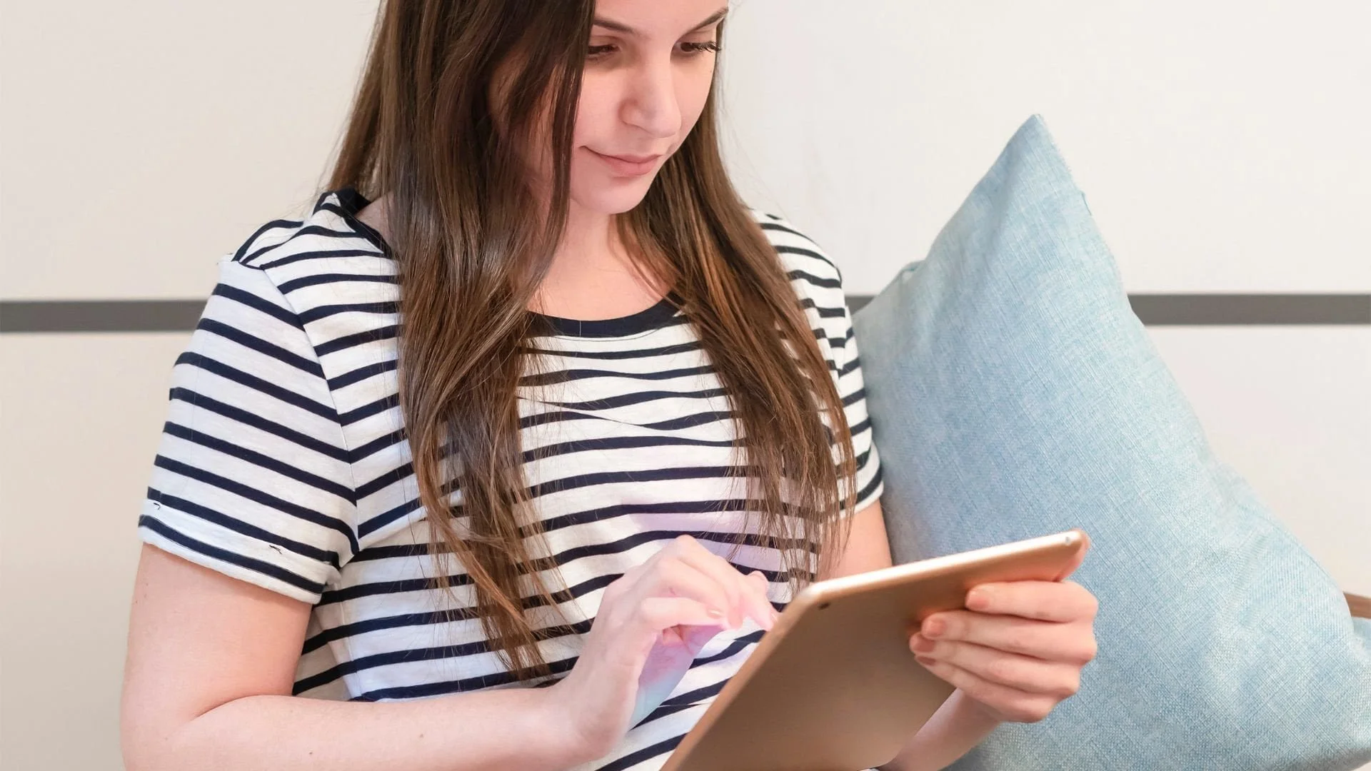Woman in a striped shirt sitting on a couch, using a tablet with one hand, with a blue pillow behind her.