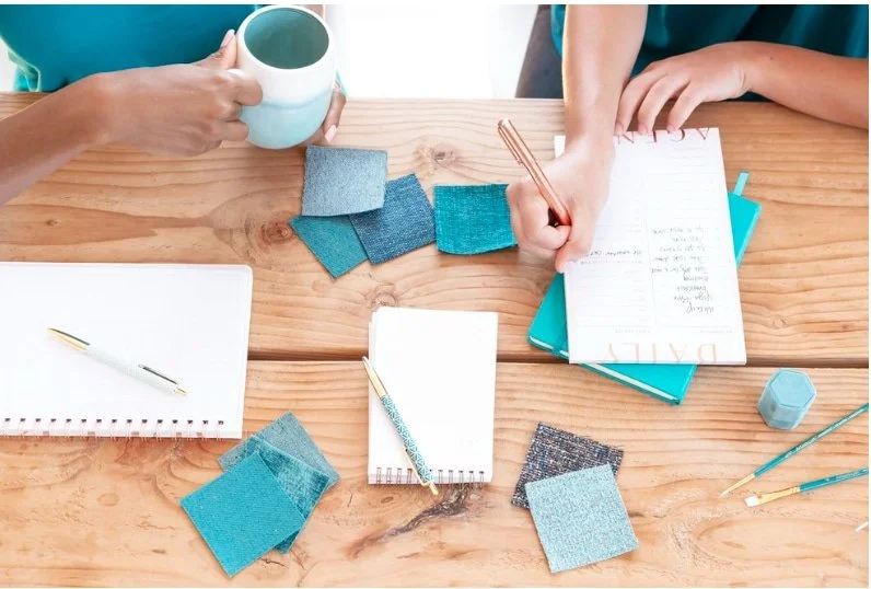 Overview of two people at a wooden table looking at aqua swatches and writing in notebooks.