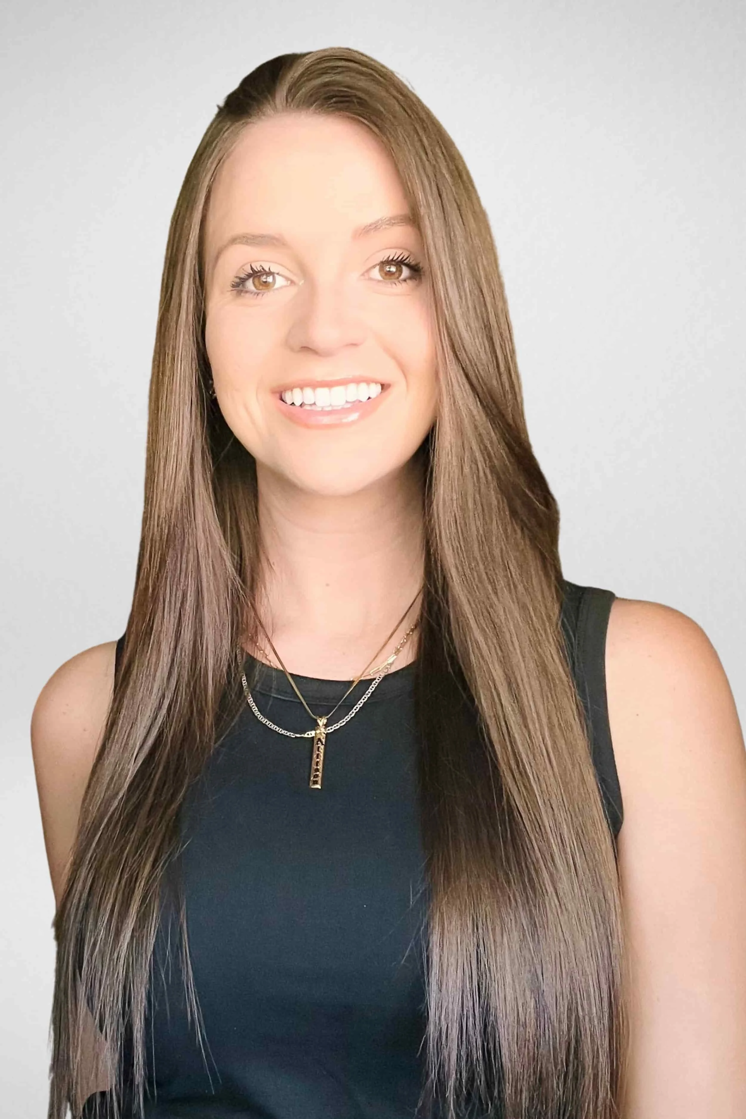 Smiling woman with long straight brown hair, wearing a black sleeveless top and layered necklaces, against a light grey background.