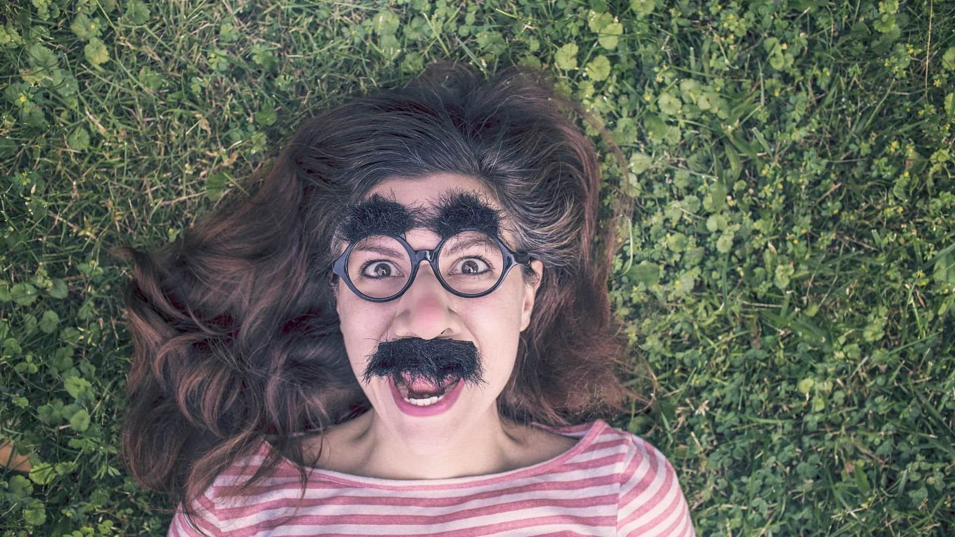Woman lying on grass, wearing a fake mustache, thick eyebrows, and glasses, making a funny surprised face.