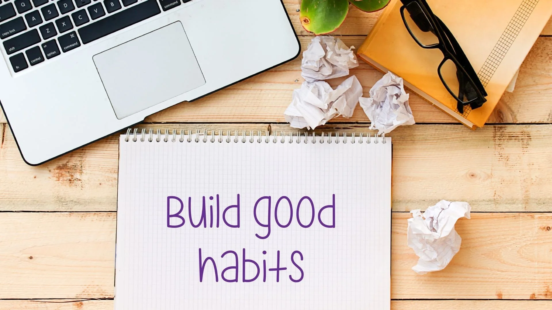 Notebook with Build good habits written on it, near a laptop, plant, glasses, and crumpled papers on a wooden desk.