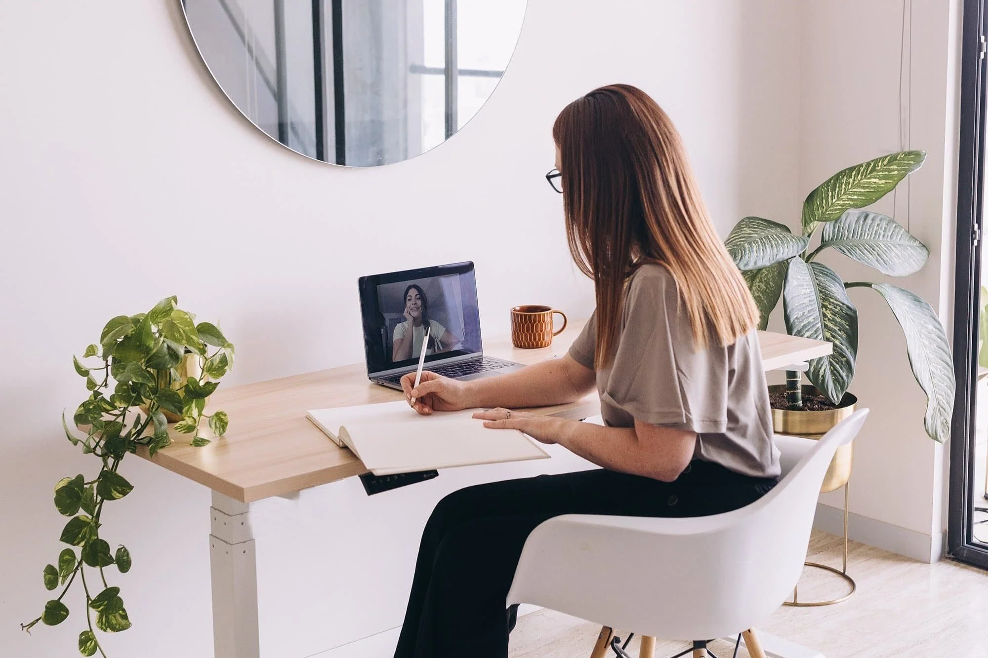 Woman with medium length light brown hair on a video meeting at a desk with plants on each side.