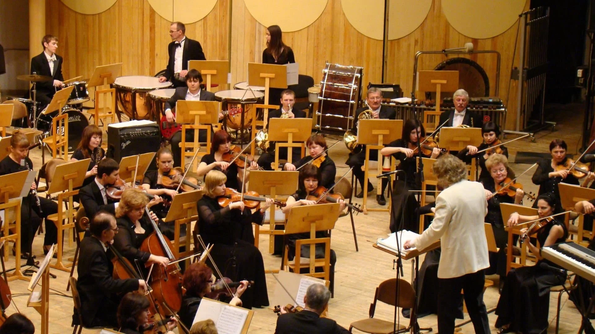 A conductor leads an orchestra of musicians playing string and percussion instruments on a wooden stage.
