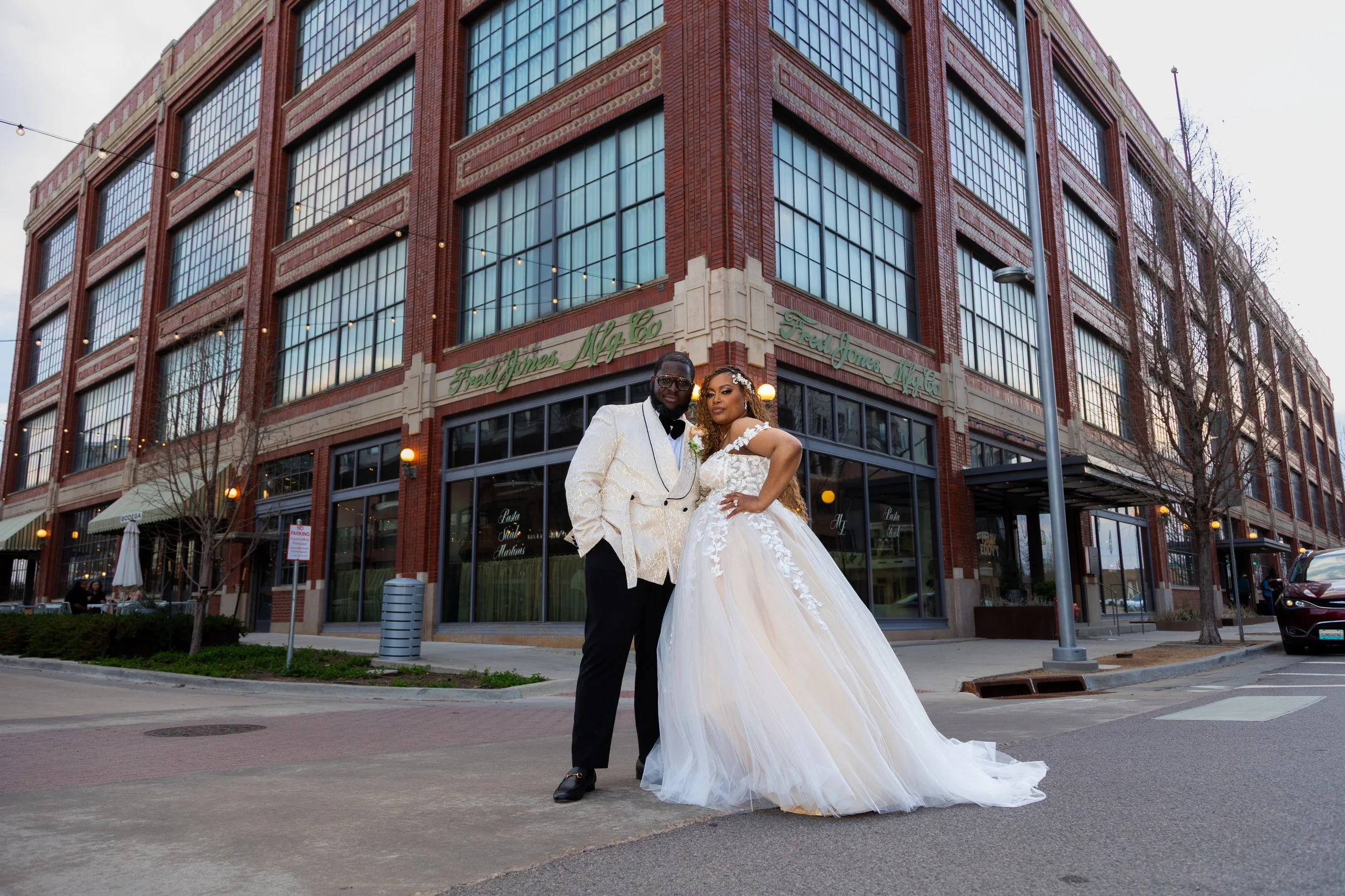 A bride and groom in wedding attire standing in Oklahoma city street in front of a brick building with large windows. The groom is wearing a white tuxedo jacket with black pants, and the bride is in a white wedding gown with floral lace details. Take