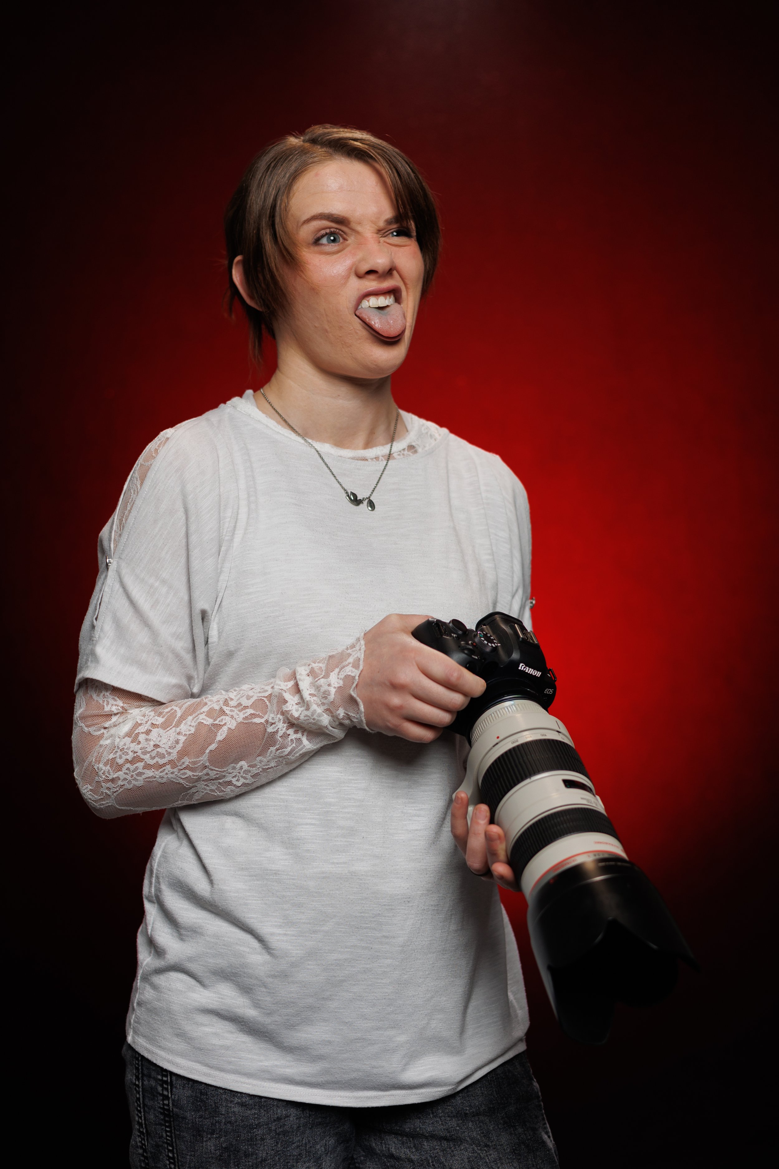 A female wedding photographer with short brown hair making a funny face and sticking out her tongue, holding a professional camera with a large zoom lens in front of a red background.