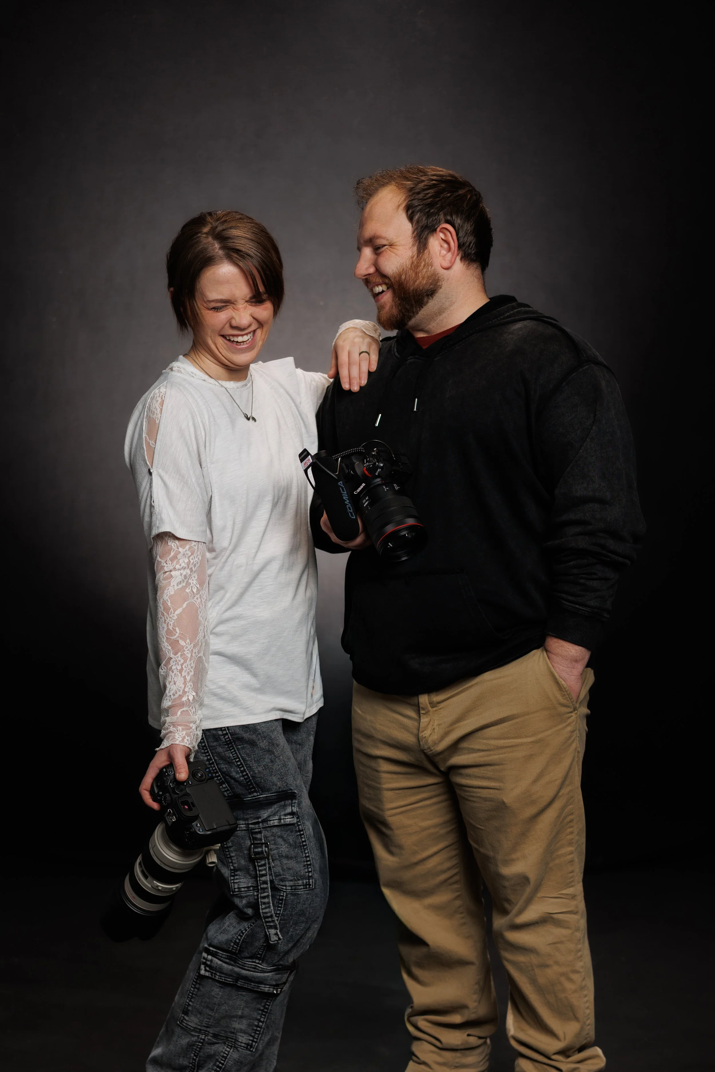 top-rated wedding photographers in Oklahoma City Two Oklahoma wedding photographers laughing and interacting against a dark backdrop, holding professional cameras.