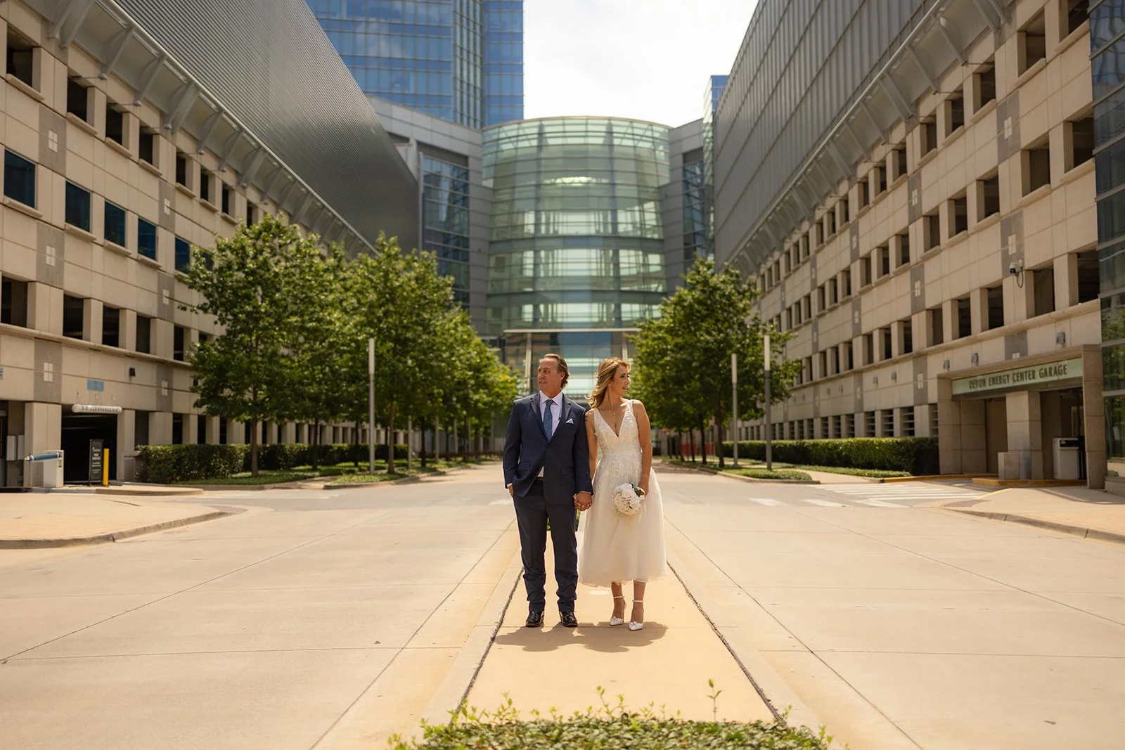 A bride and groom walking hand in hand outdoors in Oklahoma city with modern glass and concrete buildings, trees, and clear skies. Taken by top-rated wedding photographers in Oklahoma City