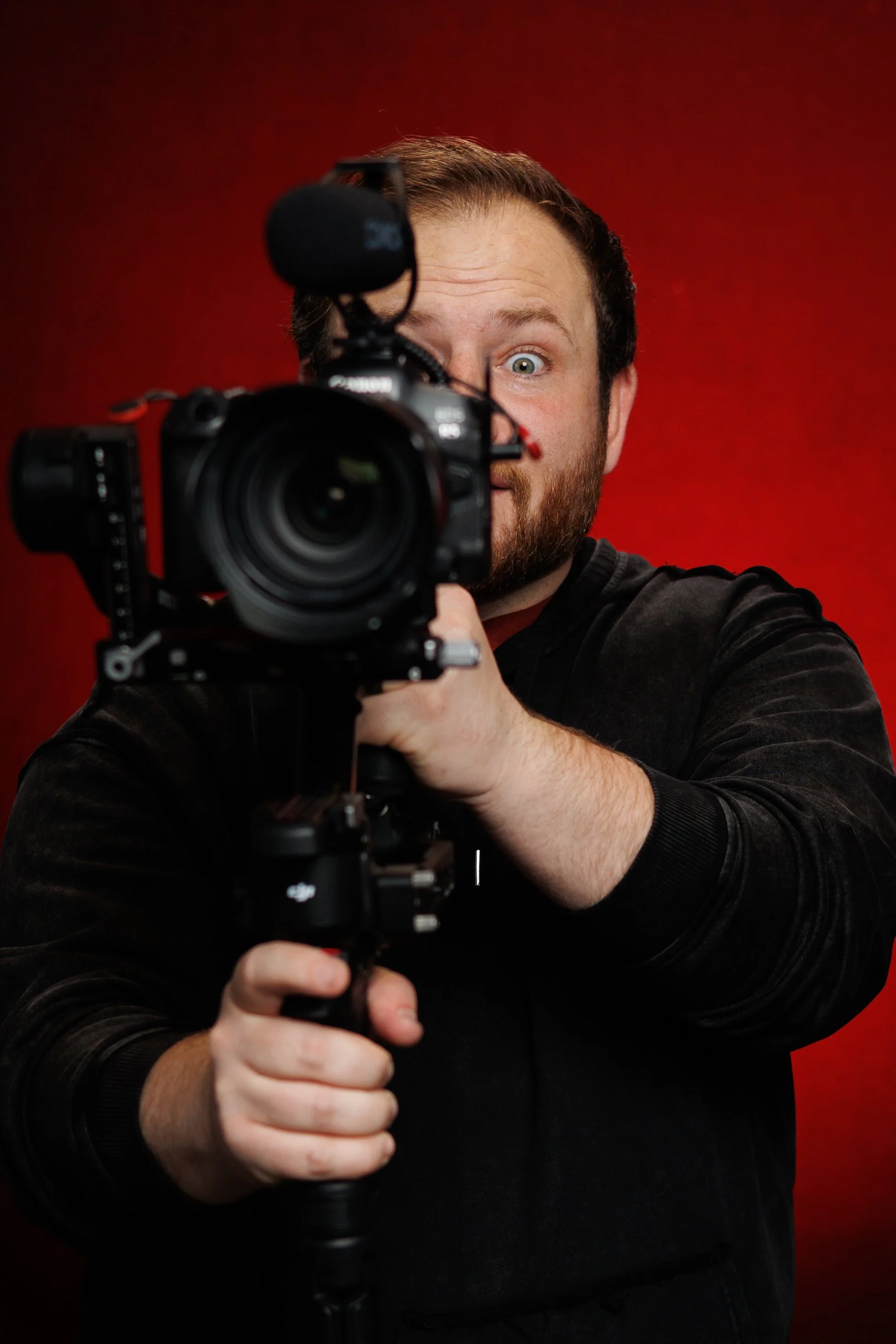 A male wedding photographer in Oklahoma city with a beard and short hair holding a professional video camera, with a surprised or intense expression, against a red background.