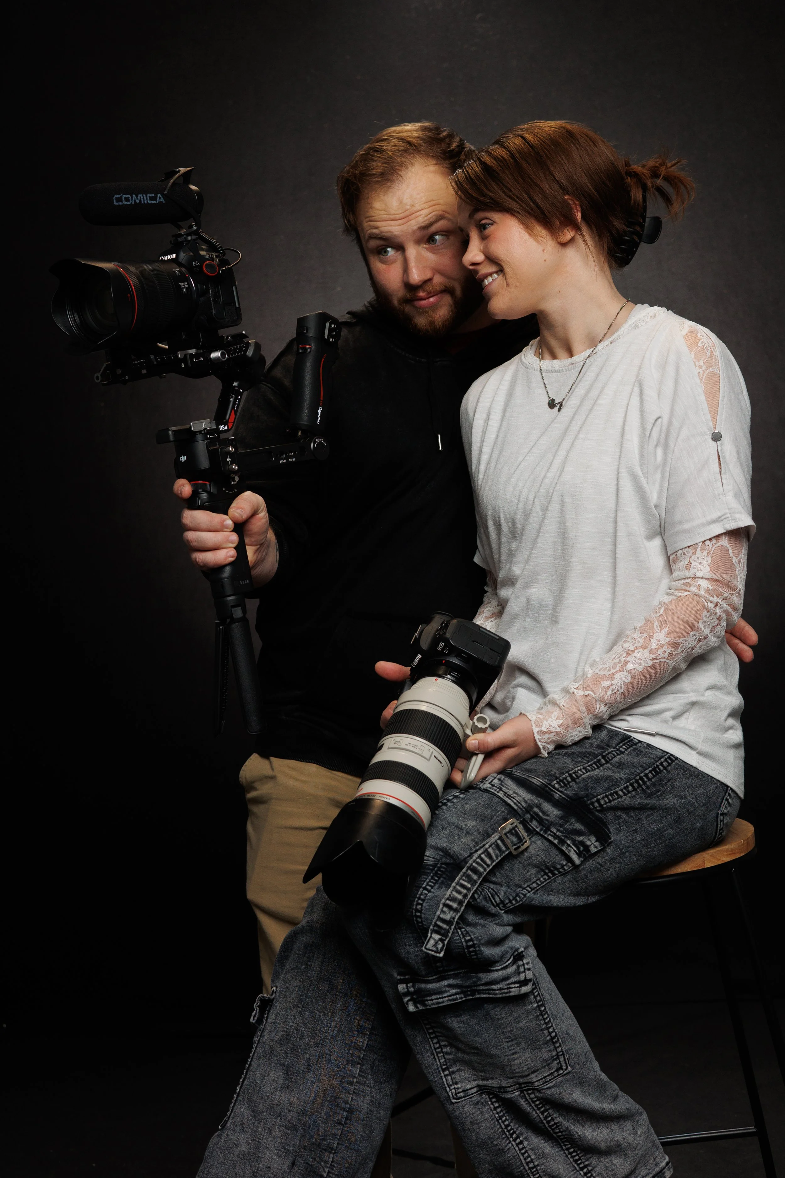 A male wedding photographer holding a professional camera with a microphone, sitting next to a female Oklahoma wedding photographer holding a camera lens, both smiling and leaning close to each other against a dark background.