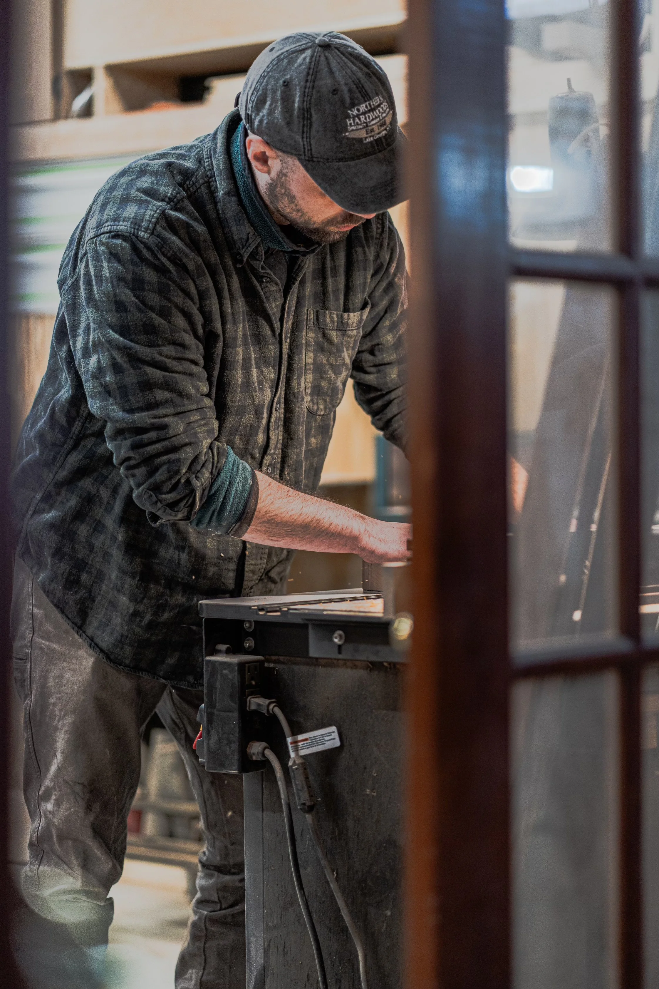A man wearing a cap and a plaid shirt working on a woodworking project in a wood shop.
