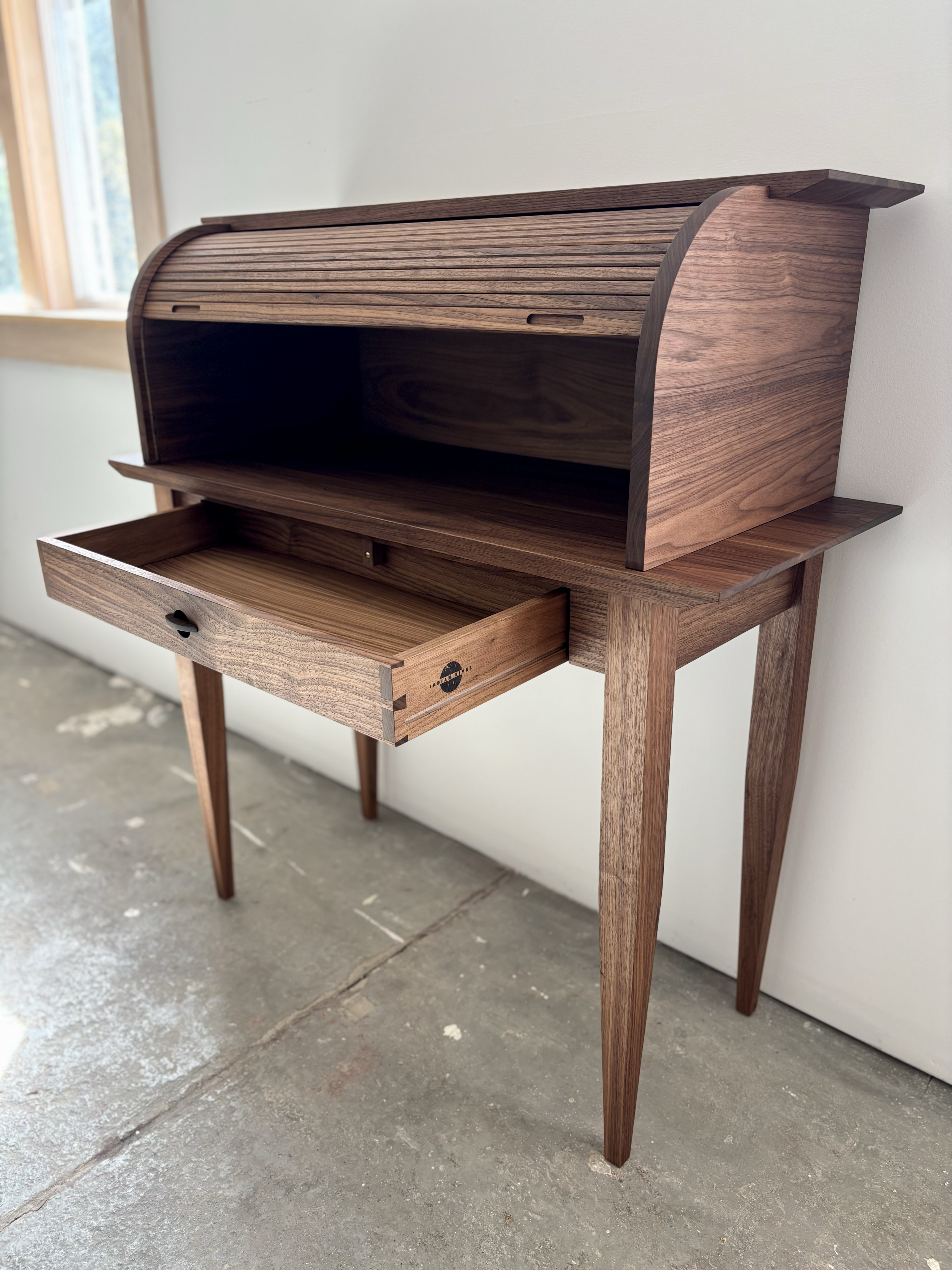 A mid-century modern wooden secretary desk with a curved roll-top, a sliding top compartment, and an open drawer, standing on four tapered legs.