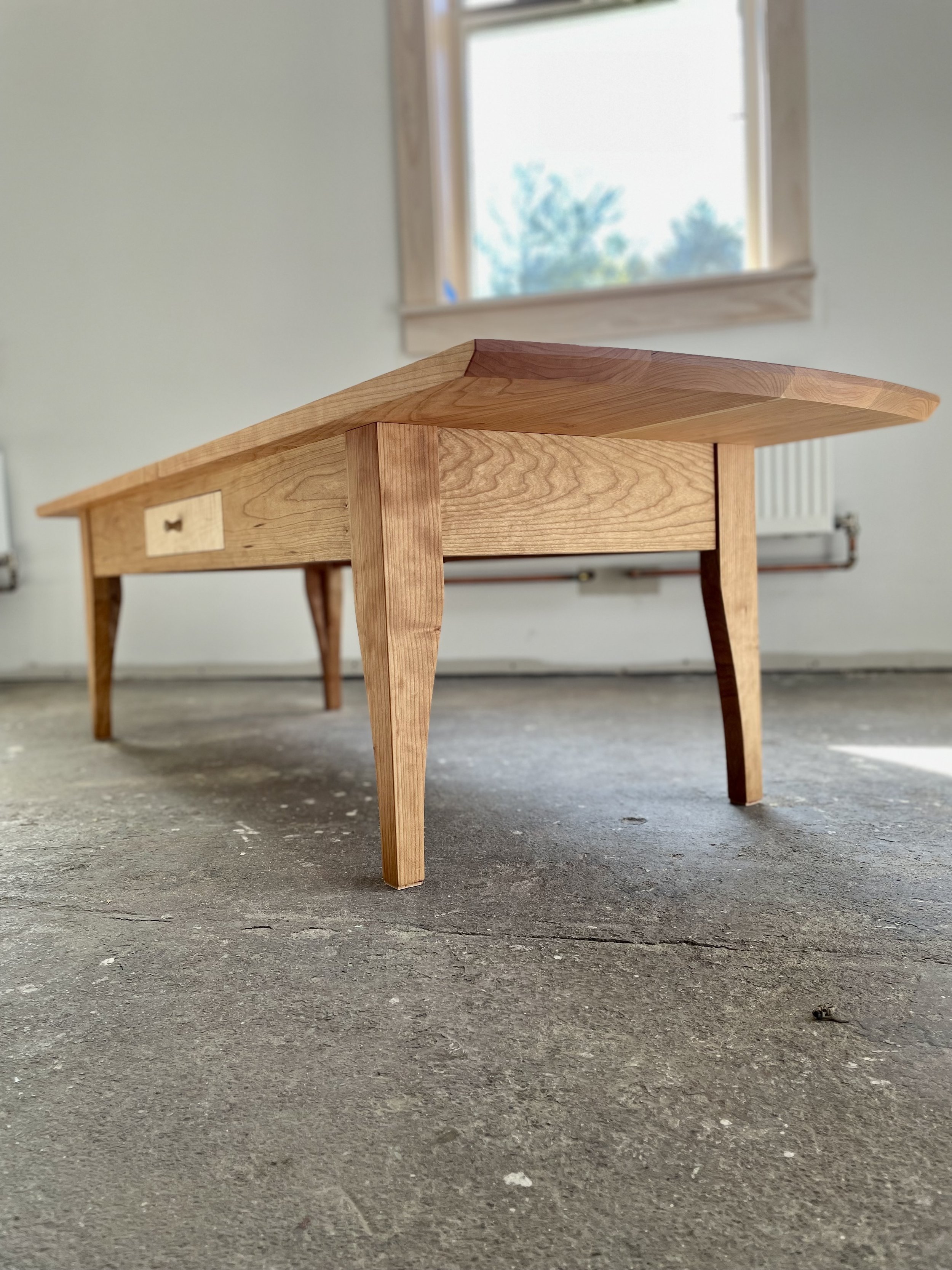Low wooden coffee table with angled legs, situated on a concrete floor near a window with wooden trim.