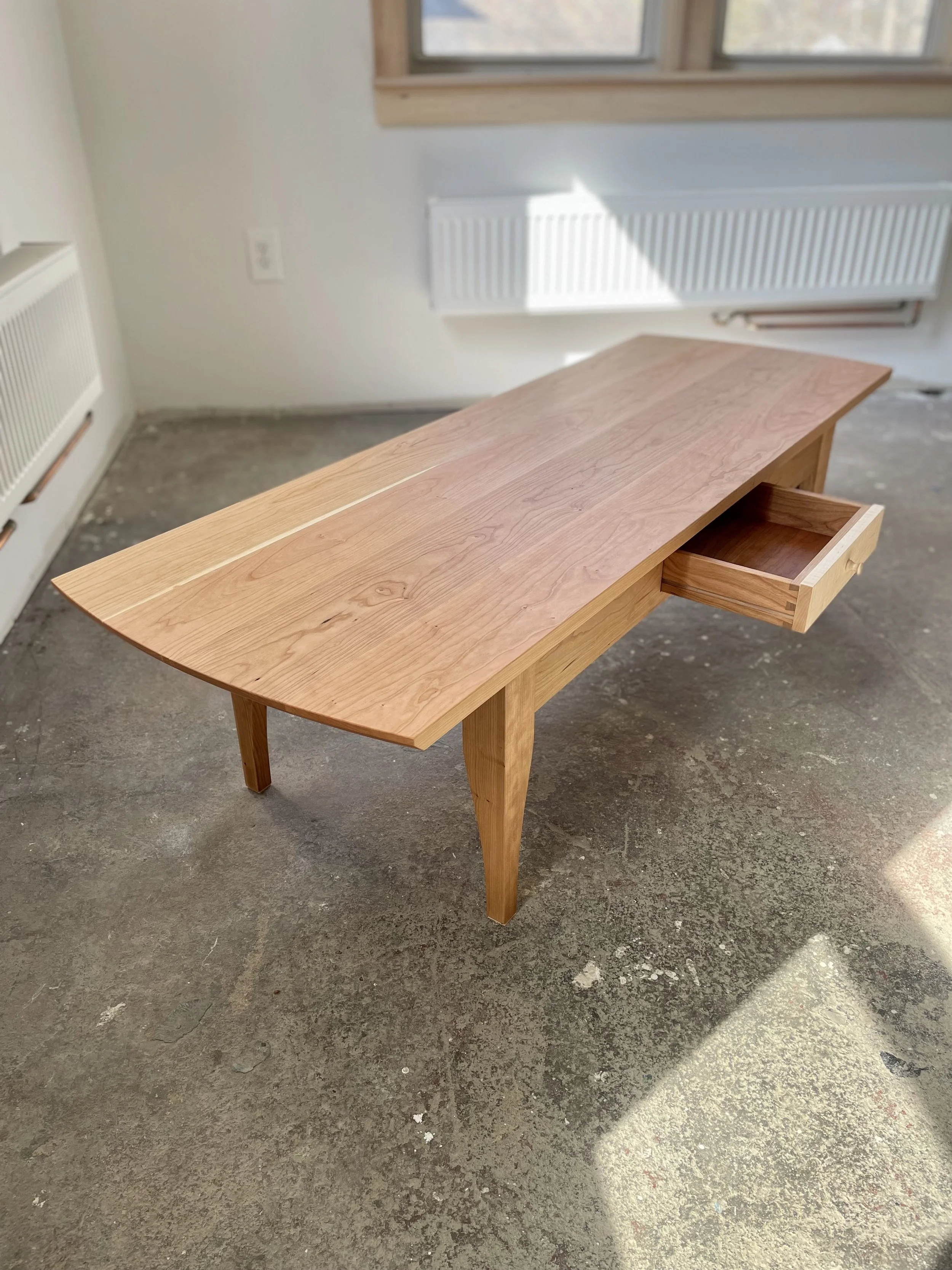 A wooden desk with a single drawer, situated on a concrete floor near a wall with a window and radiators.