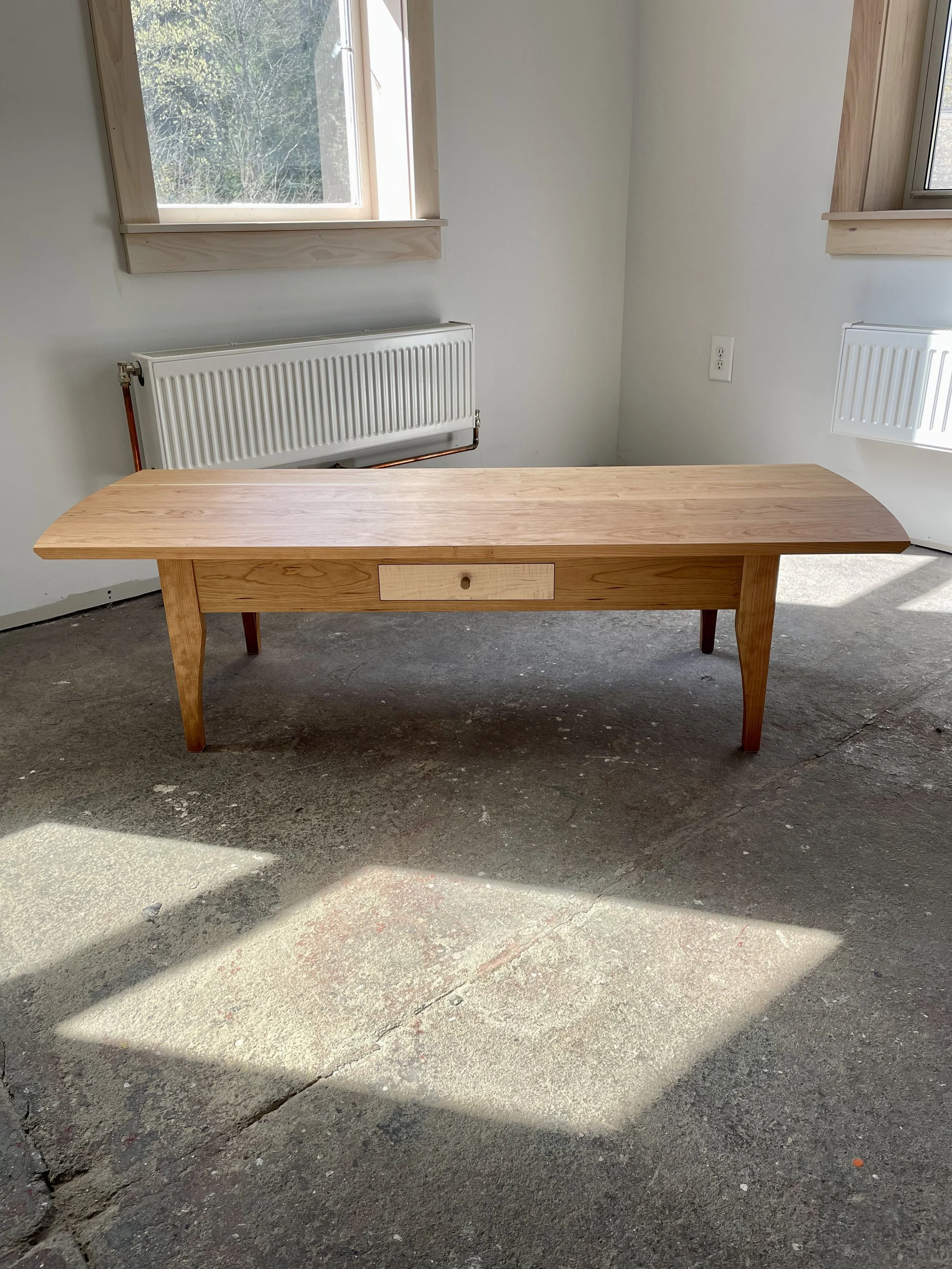 A wooden table with a single drawer in a room with concrete flooring, two windows, and a radiator underneath each window.