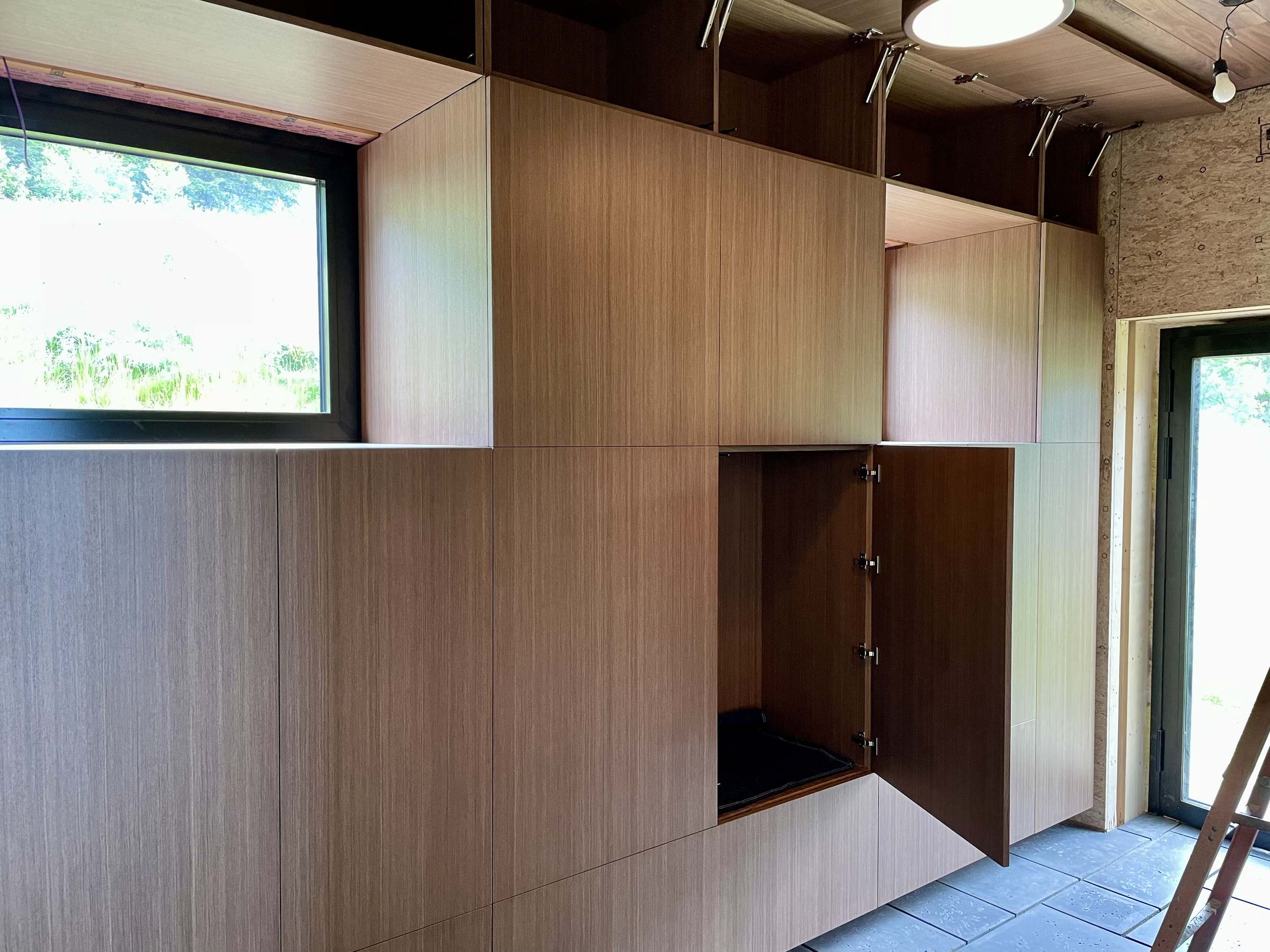 Wooden cabinets in a room under construction with open doors, windows providing natural light, and a ladder visible on the right side.