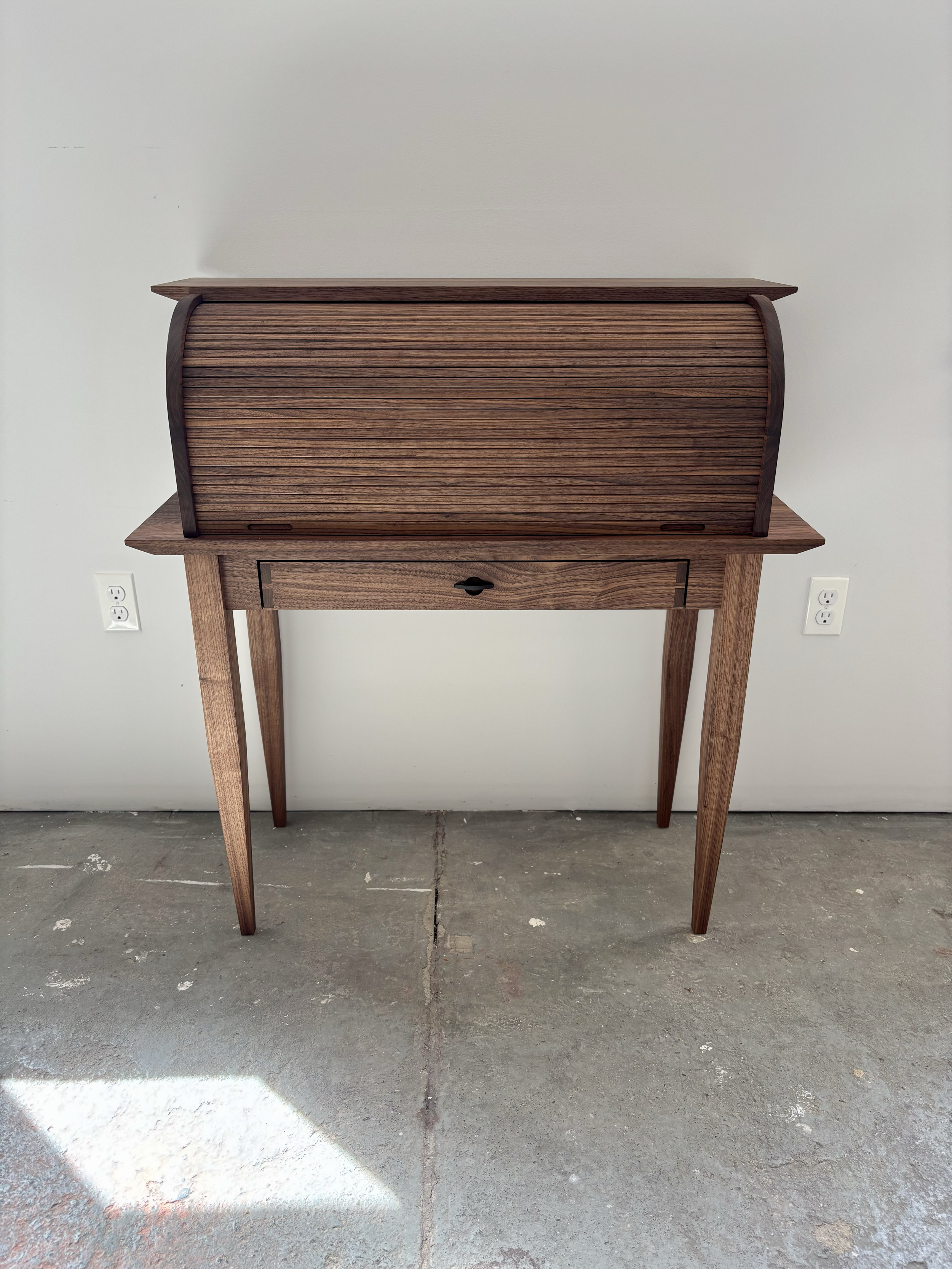 A vintage wooden secretary desk with a slanted roll-top, a small drawer beneath, and four tapered legs, placed against a white wall with electrical outlets.