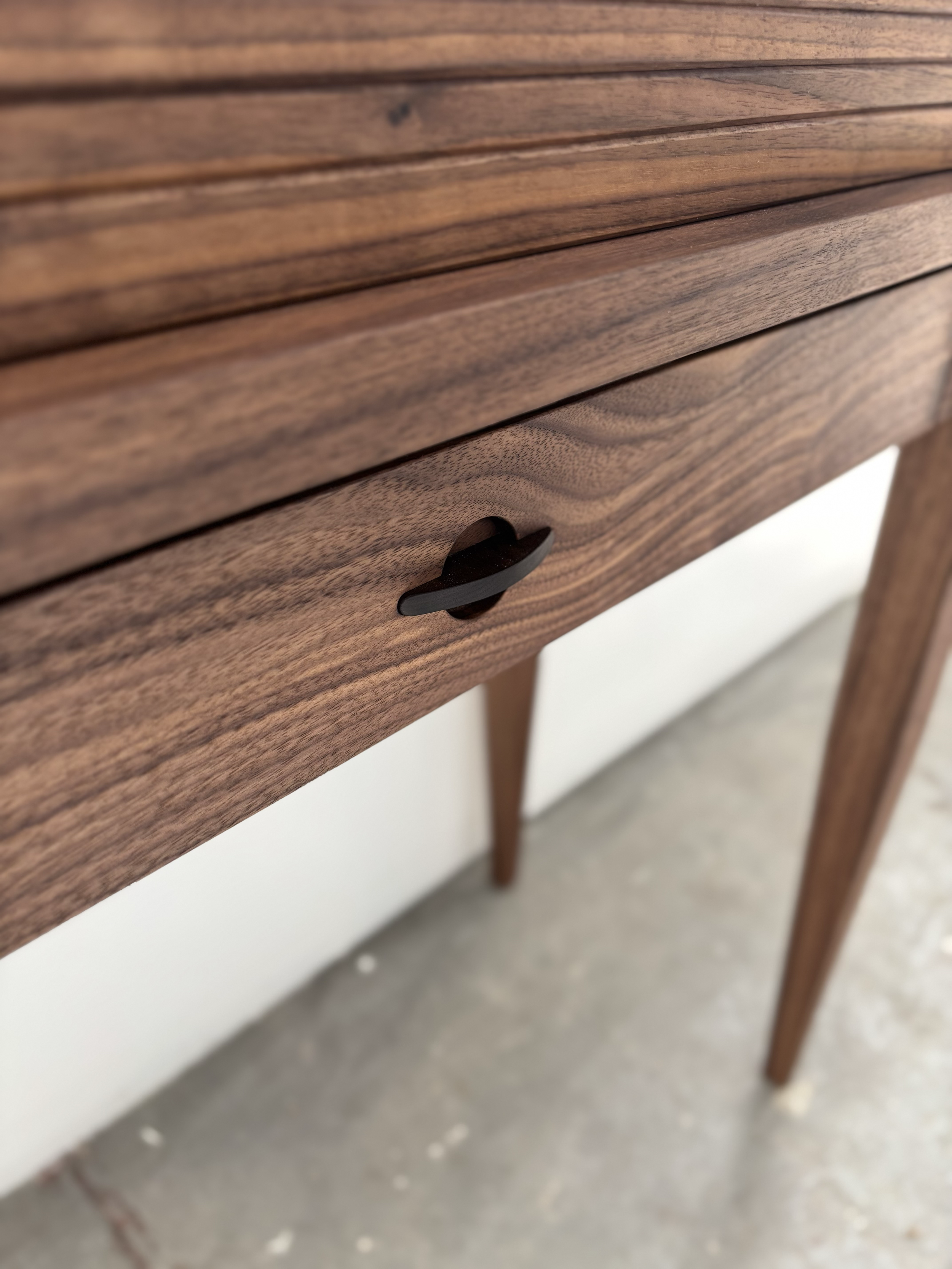 Close-up of a wooden piece of furniture with a dark handle on a drawer, showing the grain and texture of the wood.