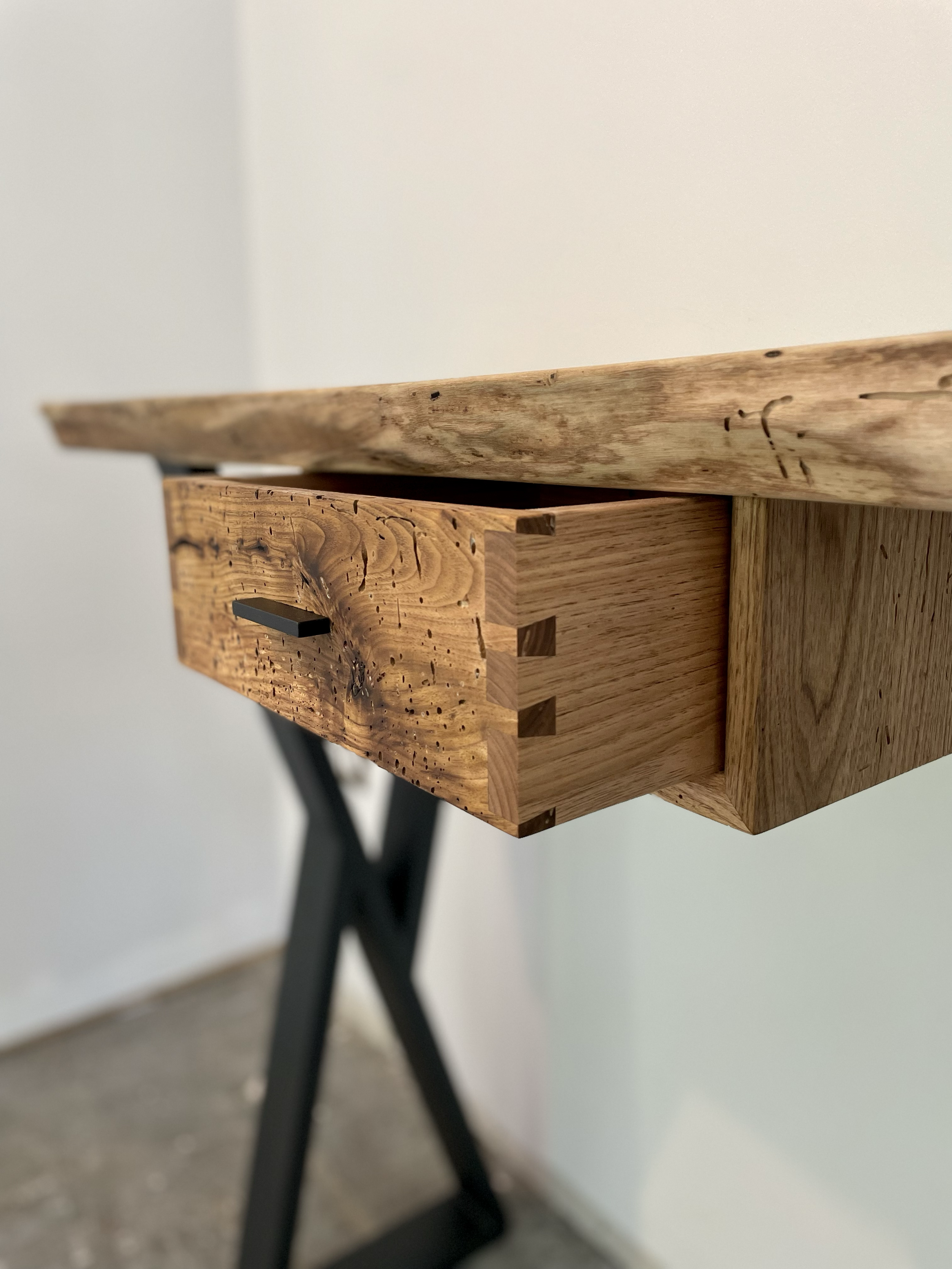 Close-up of a wooden desk with a left side drawer, showing dovetail joints and a black handle, supported by black crossed legs, against plain wall background.
