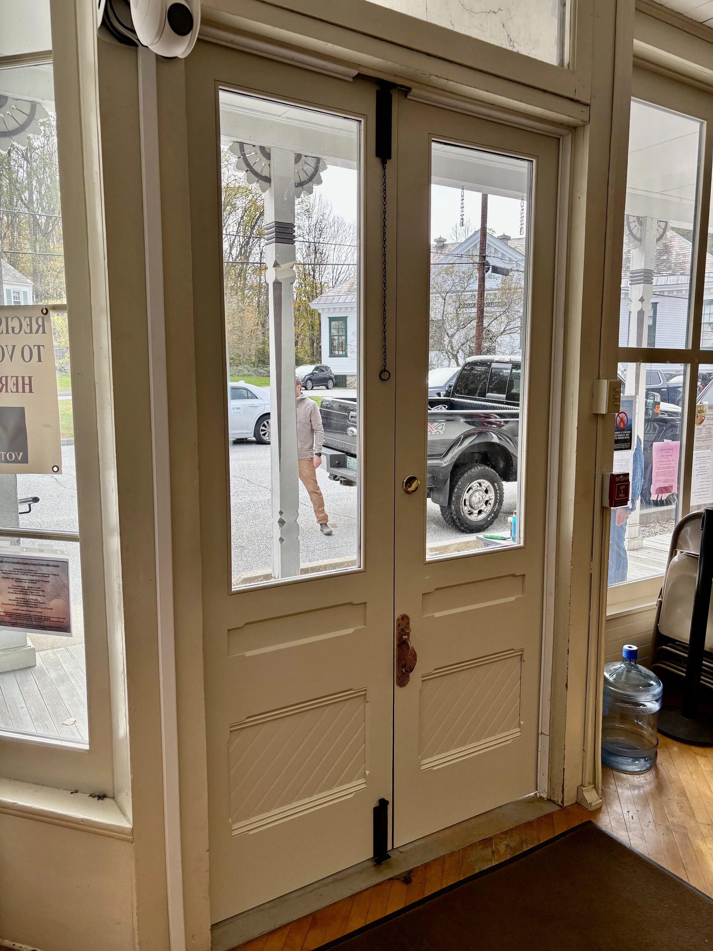 View of the front door of a building from inside, showing the glass panels, door handle, and nearby objects including a large water bottle, chairs, and notices on the door, with people and parked cars visible outside.