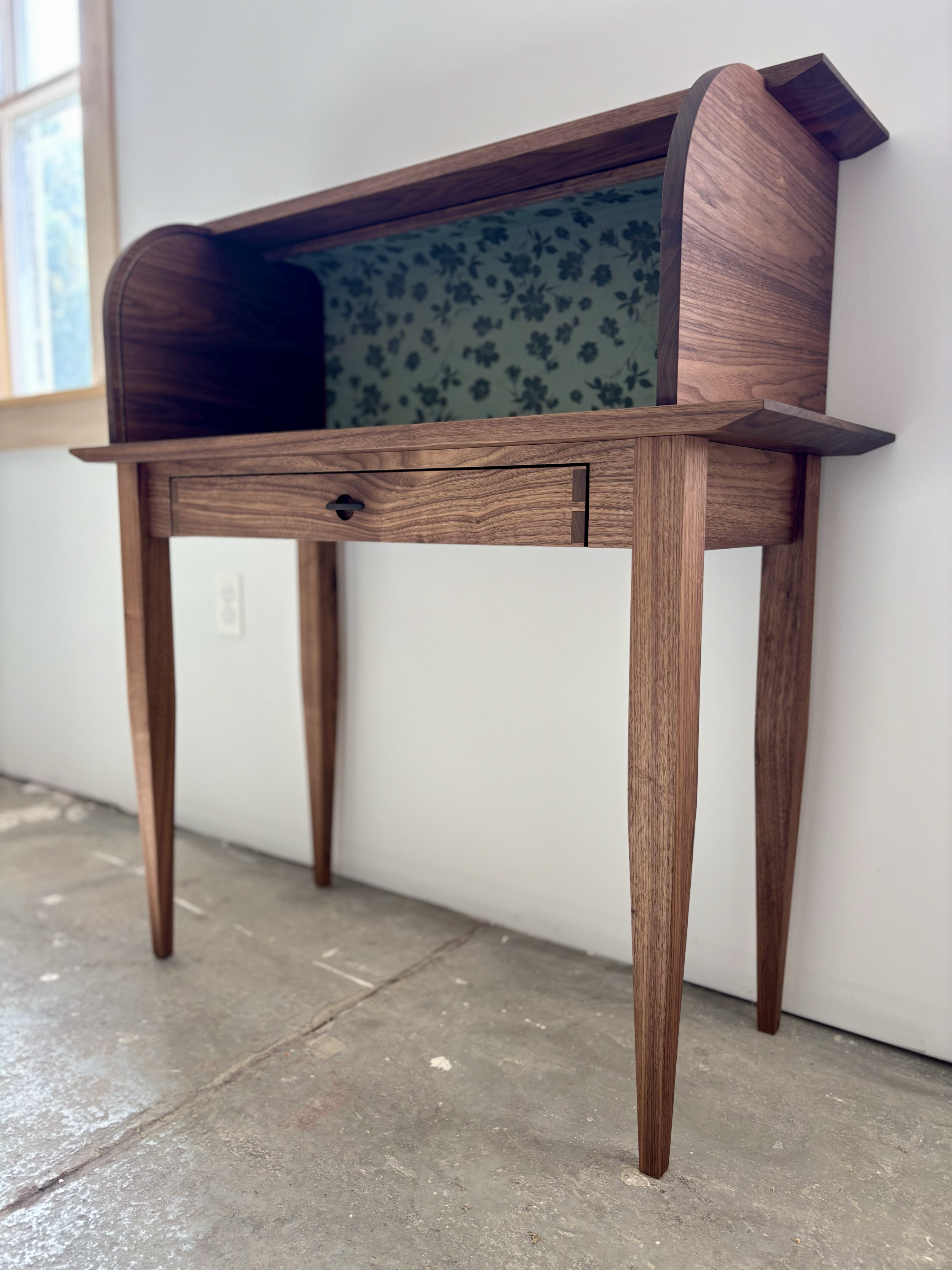 A vintage wooden desk or vanity with an open shelf space and a small drawer, featuring a floral-patterned interior lining, placed on a concrete floor near a window.