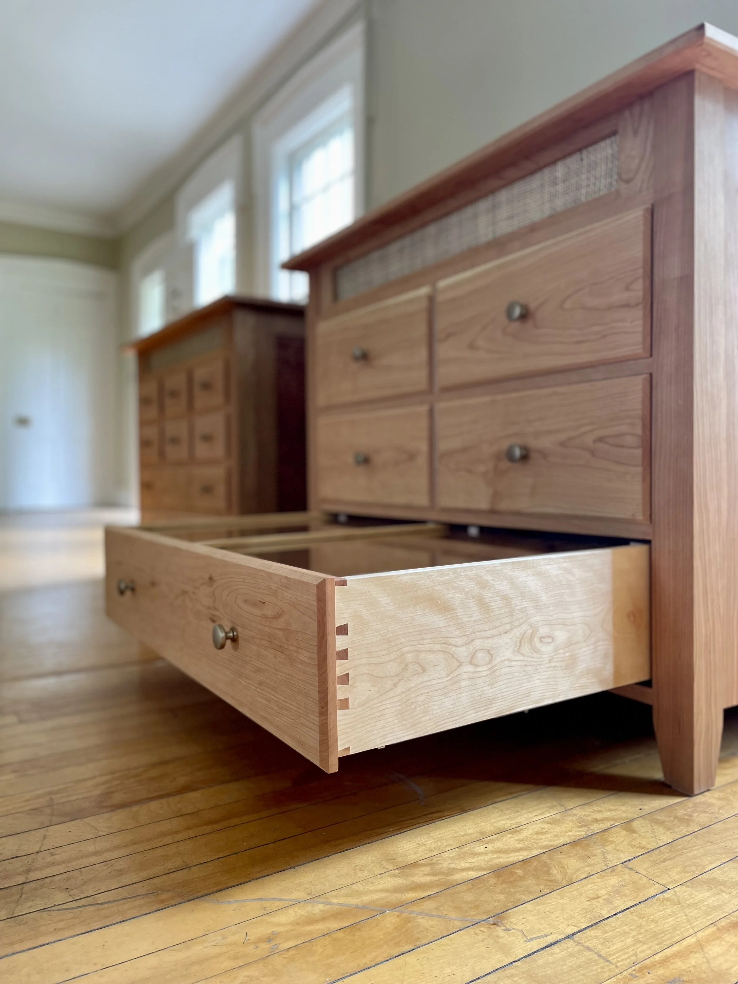 A partially pulled out wooden drawer in a wooden dresser, showcasing dovetail joints, in a room with hardwood floor and multiple windows.
