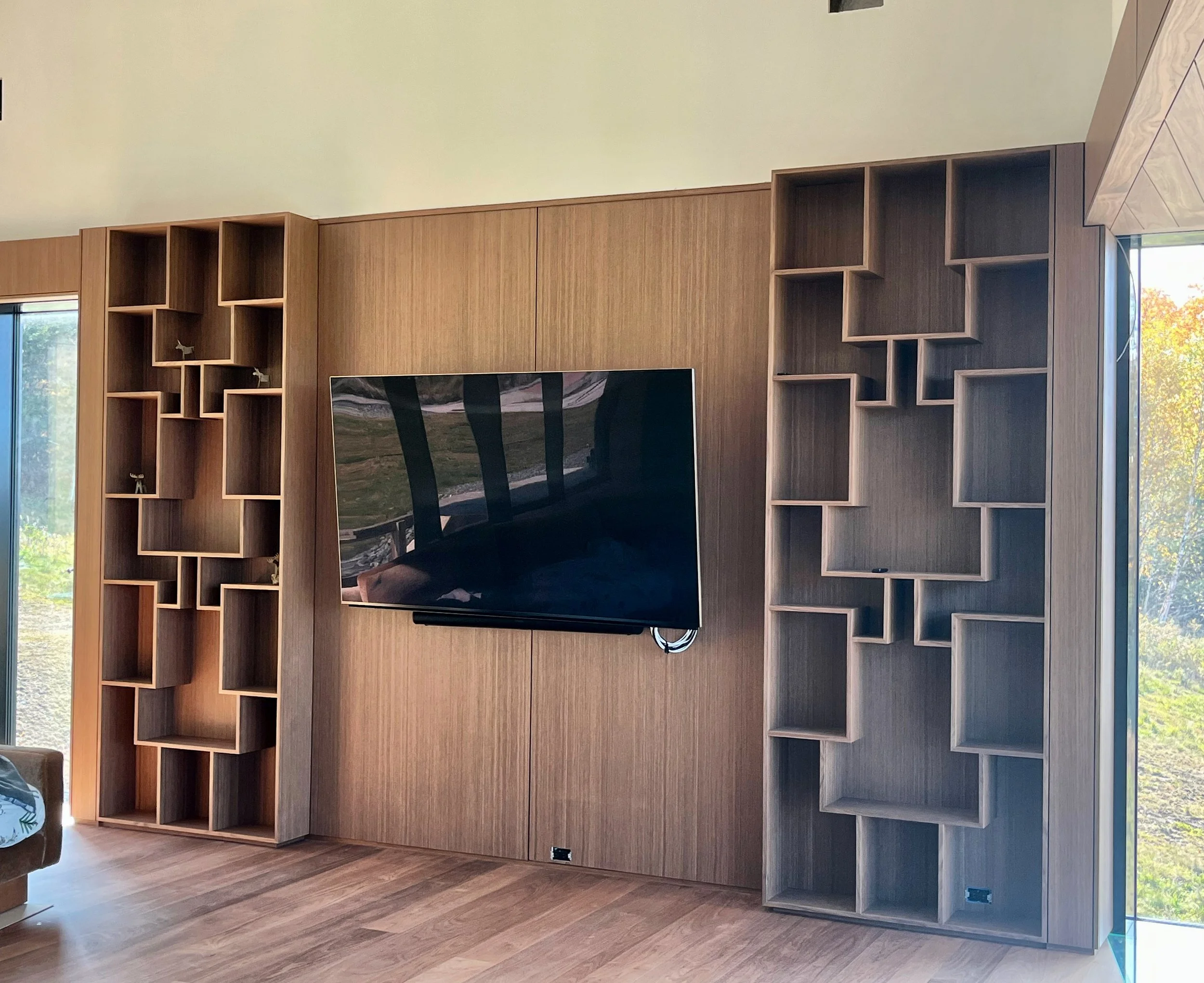Modern entertainment room with a mounted flat-screen TV on a wood-paneled wall, flanked by open shelving units with a geometric design, and large windows showing outdoor greenery.