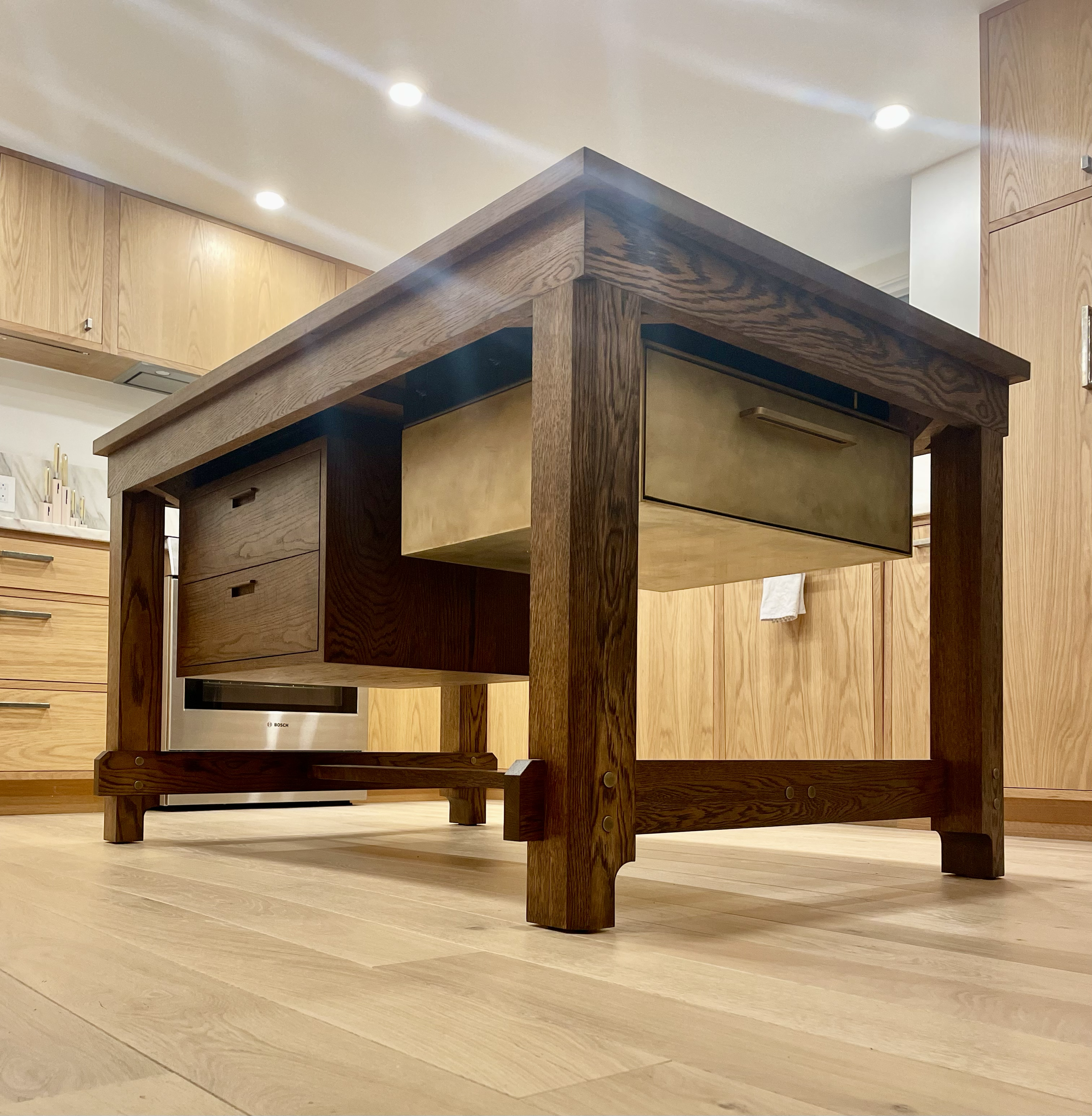 A wooden kitchen island with storage drawers and a brass-colored drawer, set in a kitchen with light wood cabinetry and flooring.