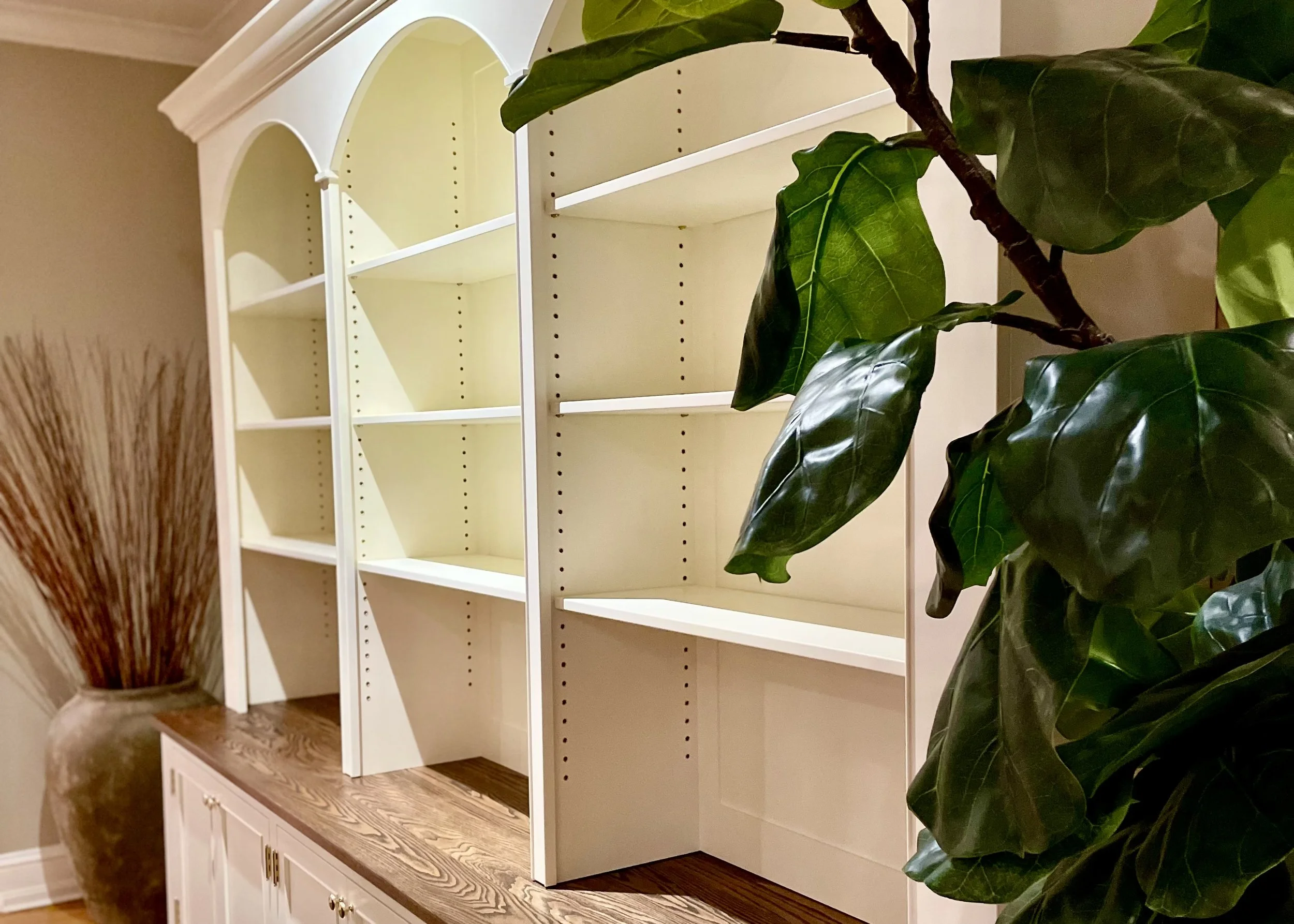 Empty white bookshelf with three sections, section on left has an arched top, on a wooden cabinet with leaf-shaped door handles, next to green leafy plant and a large vase with dried branches.