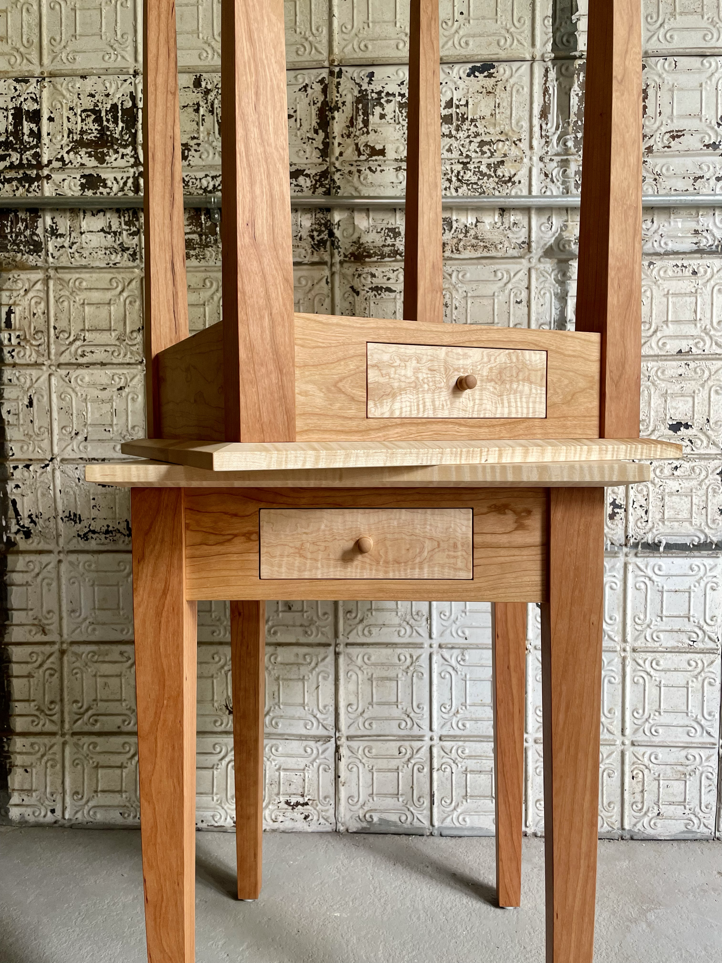 A wooden furniture piece with three tiers, each with a small drawer and knob, against a vintage tin ceiling wall. The piece is minimally designed, featuring clean lines and a natural wood finish.