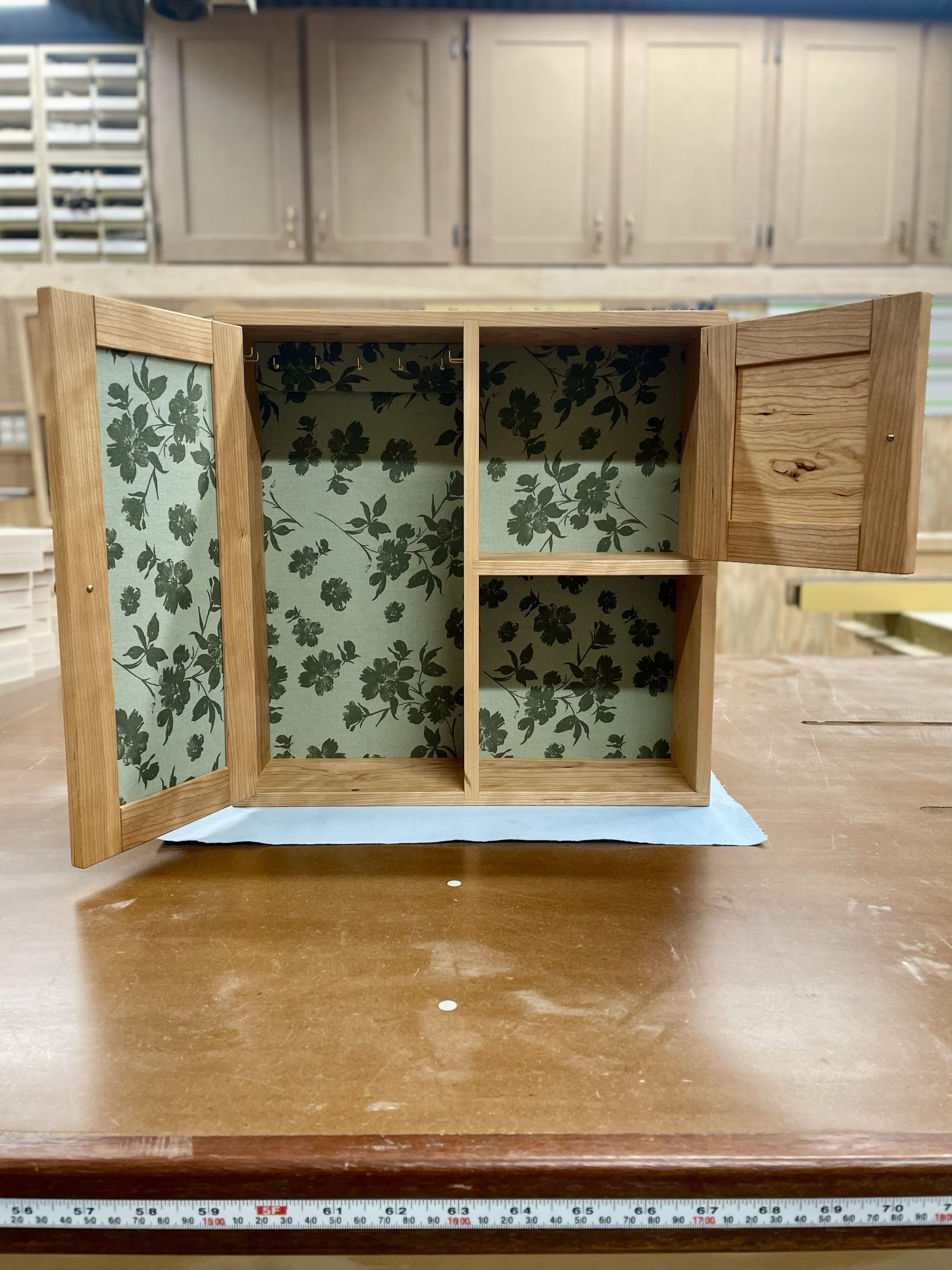 A small wooden cabinet with floral-patterned wallpaper on the interior, placed on a wooden table in a workshop.