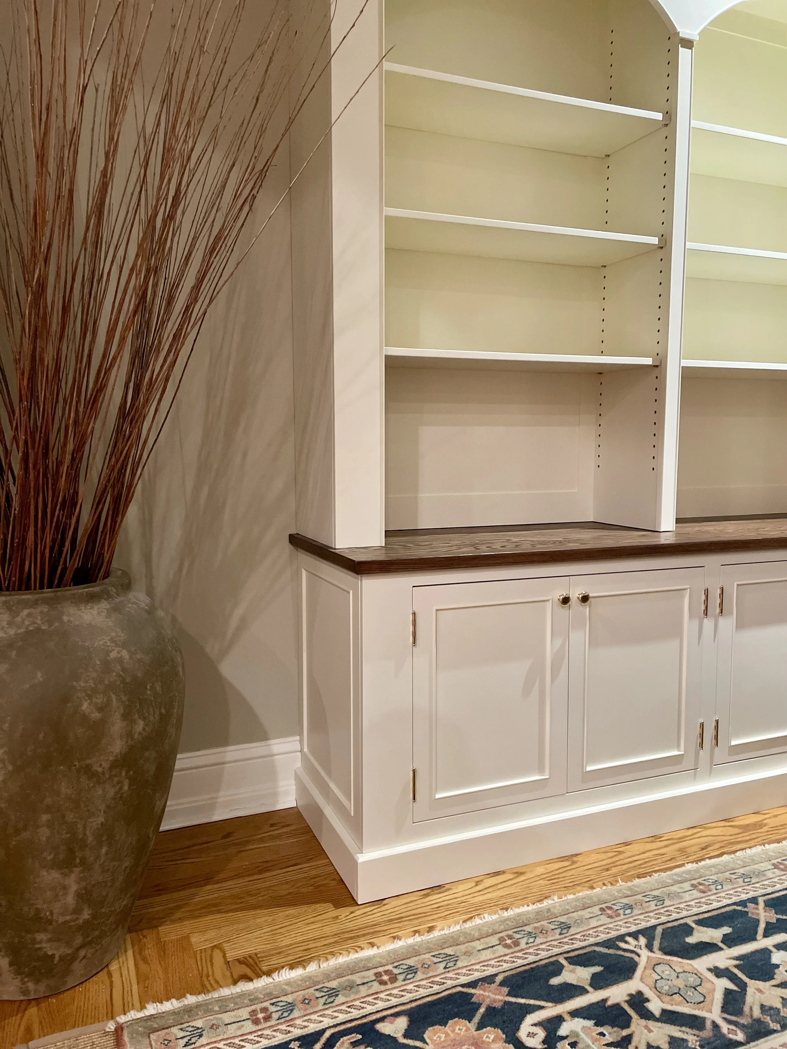 A white built-in cabinet with open shelves and closed storage, decorated with a dark wood countertop. Beside it is a large vase with tall, dried grass and a patterned area rug on wooden floor.