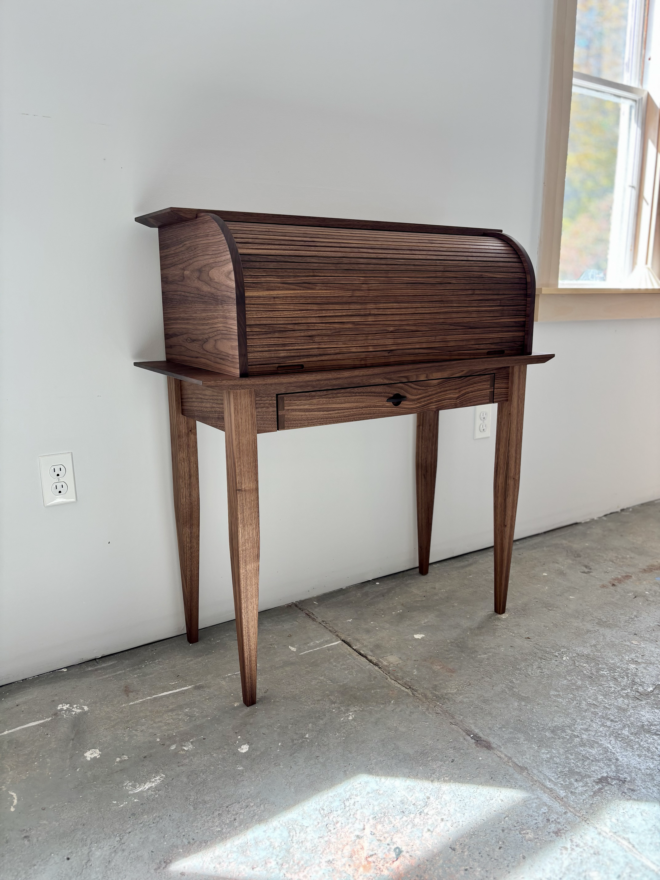 A vintage wooden writing desk with a curved roll-top lid, a small drawer underneath, and four tapered legs, placed against a plain wall next to a window.