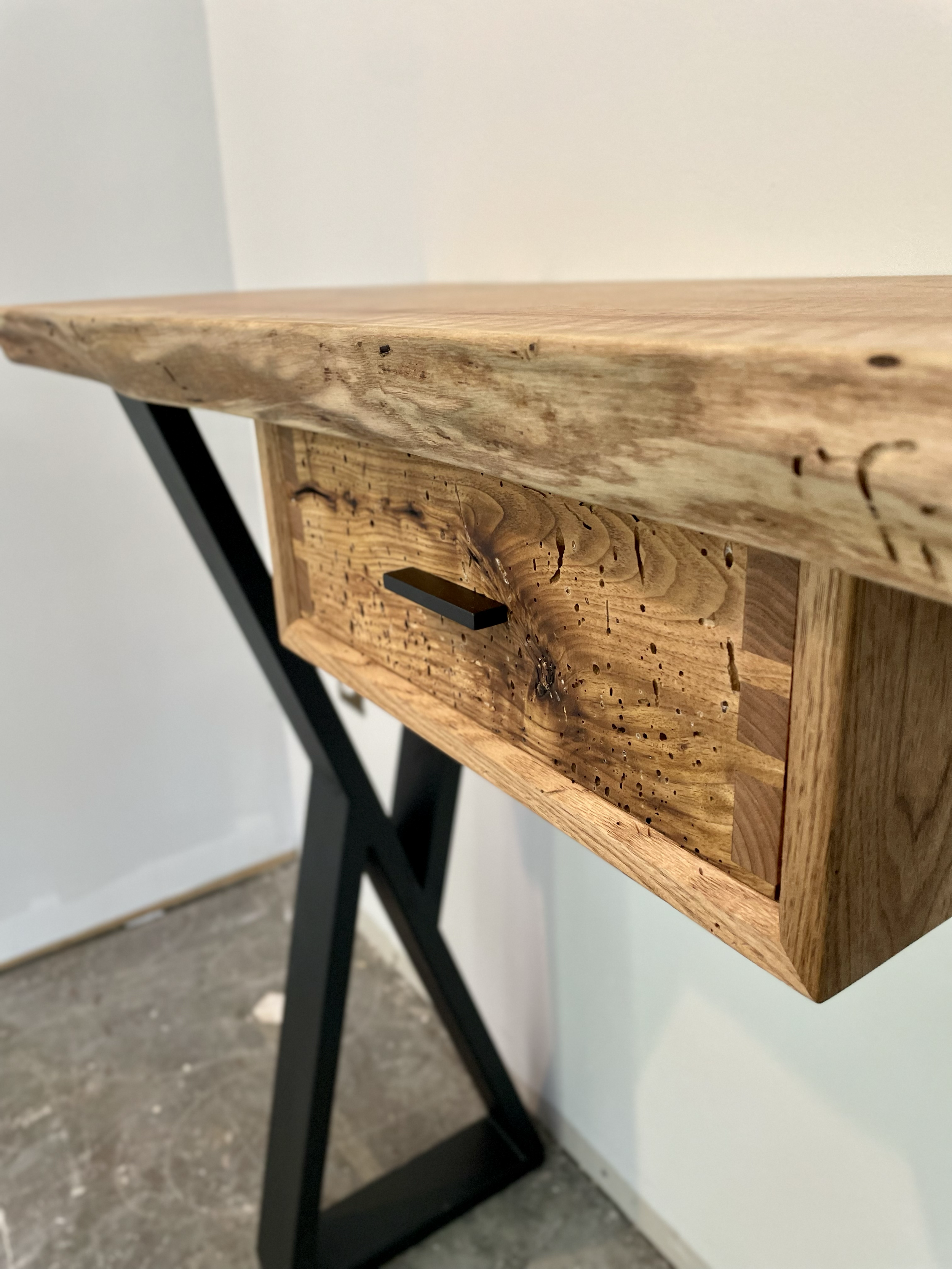 Close-up of a wooden furniture piece with natural wood grain, supported by black metal legs, set against a plain white wall.
