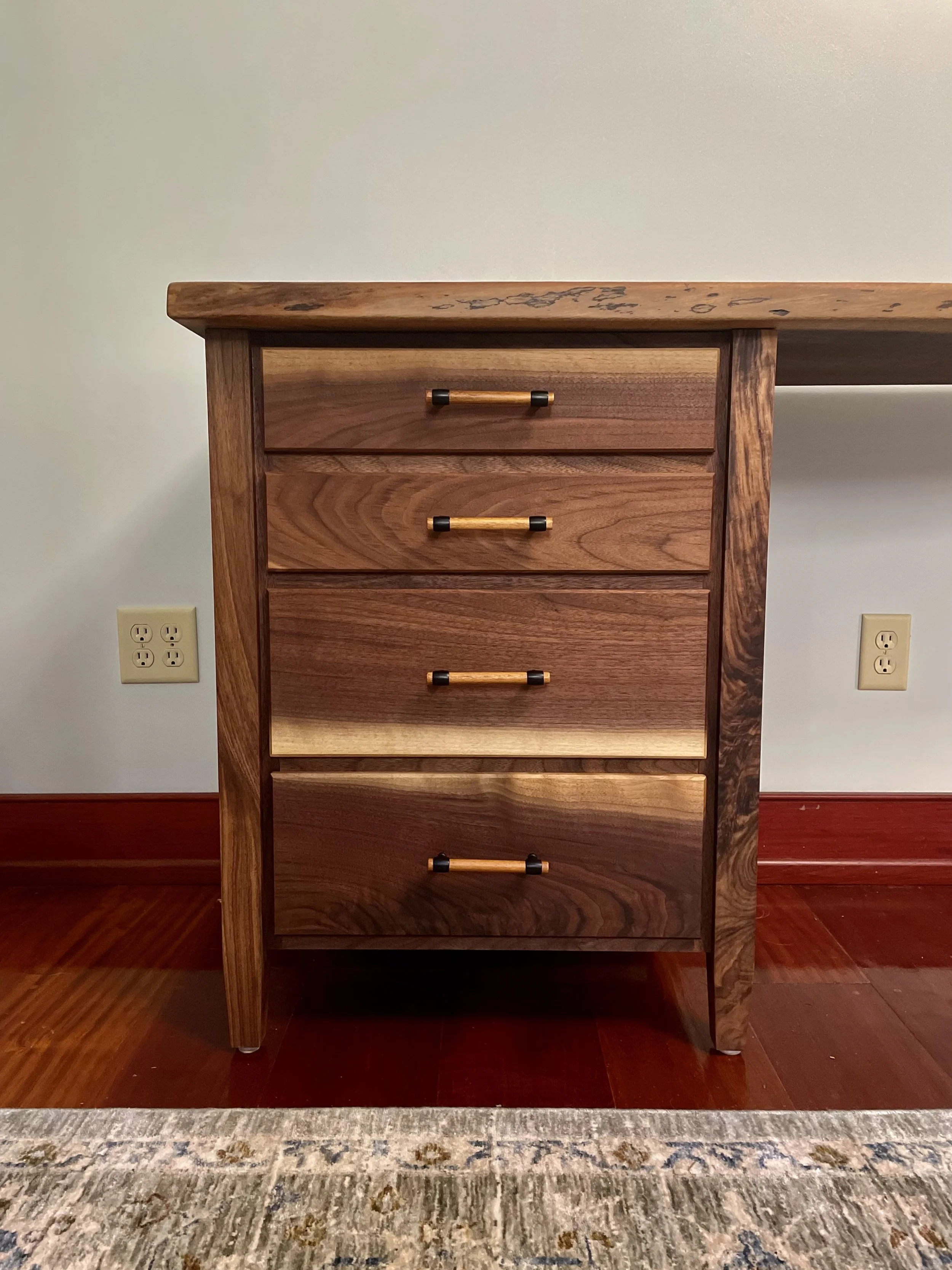 A wooden dresser with four drawers, each with a horizontal handle, standing on a hardwood floor near a white wall with two electrical outlets.