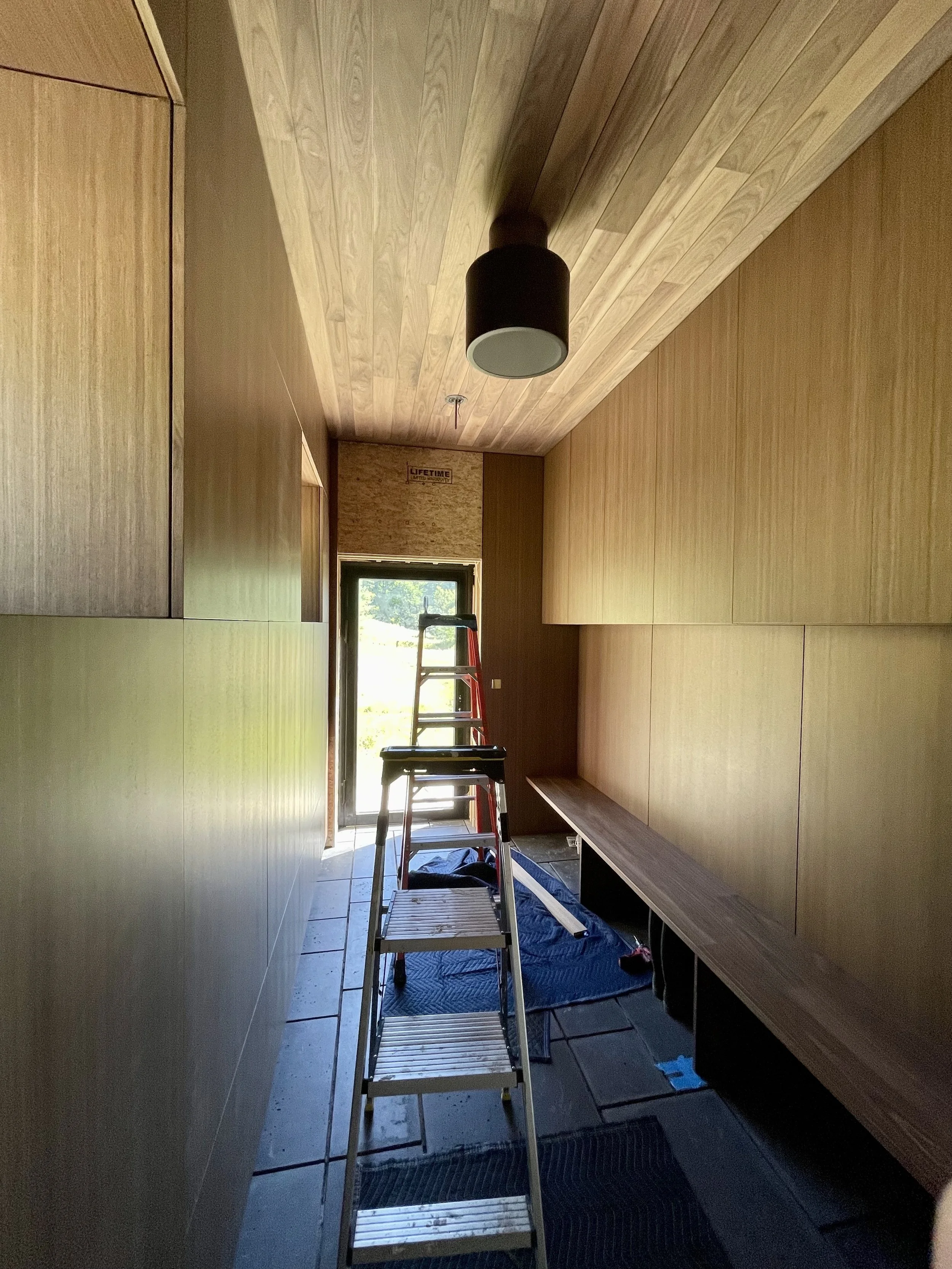 Interior view of a narrow room with wood-paneled walls and ceiling, a black ceiling light fixture, a ladder, and a wooden bench along the wall, with a door at the end leading outside.