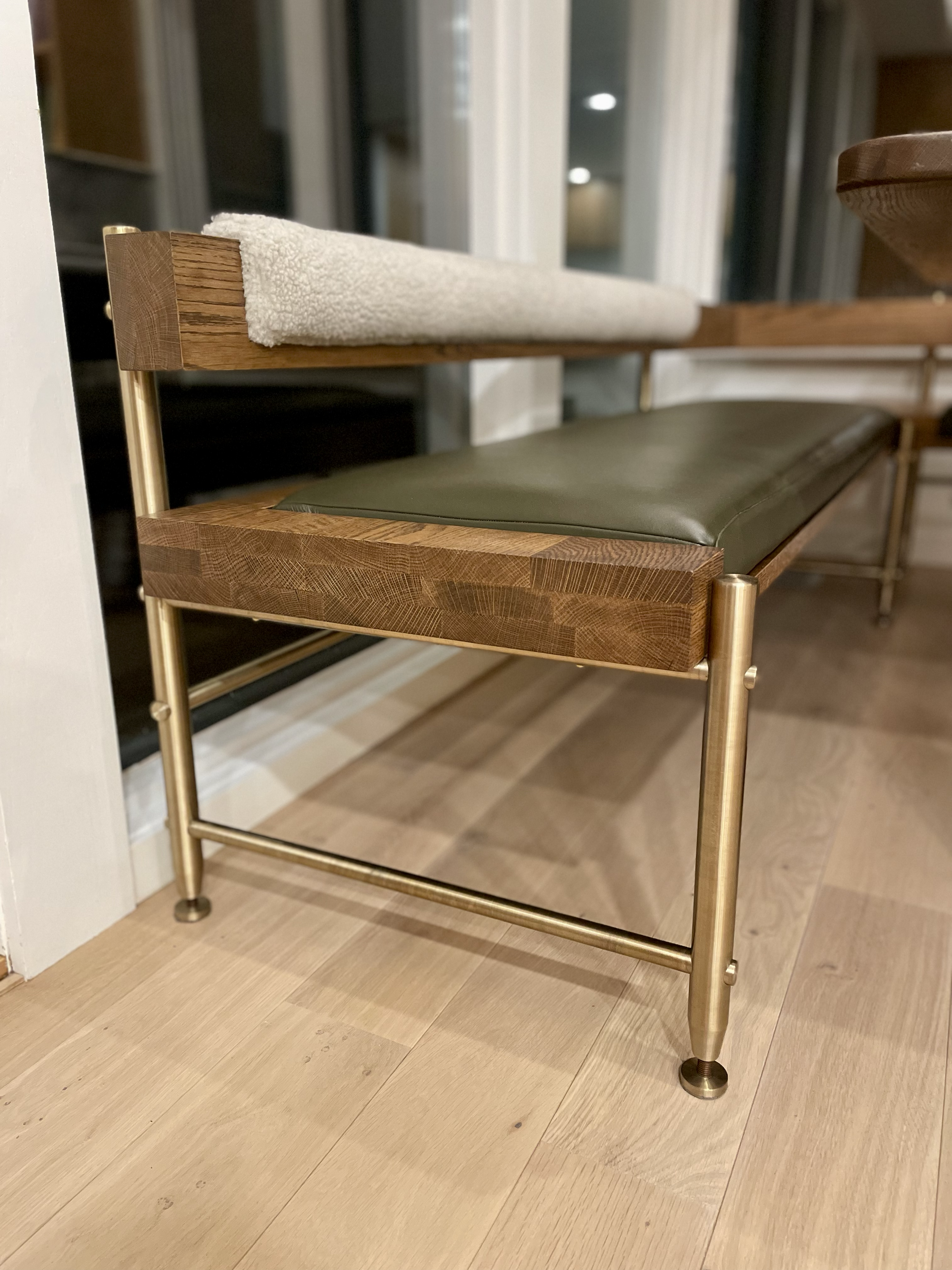 A modern bench with a wooden frame, a green cushioned seat, and a white towel draped over the backrest, located indoors near a glass sliding door and wooden flooring.